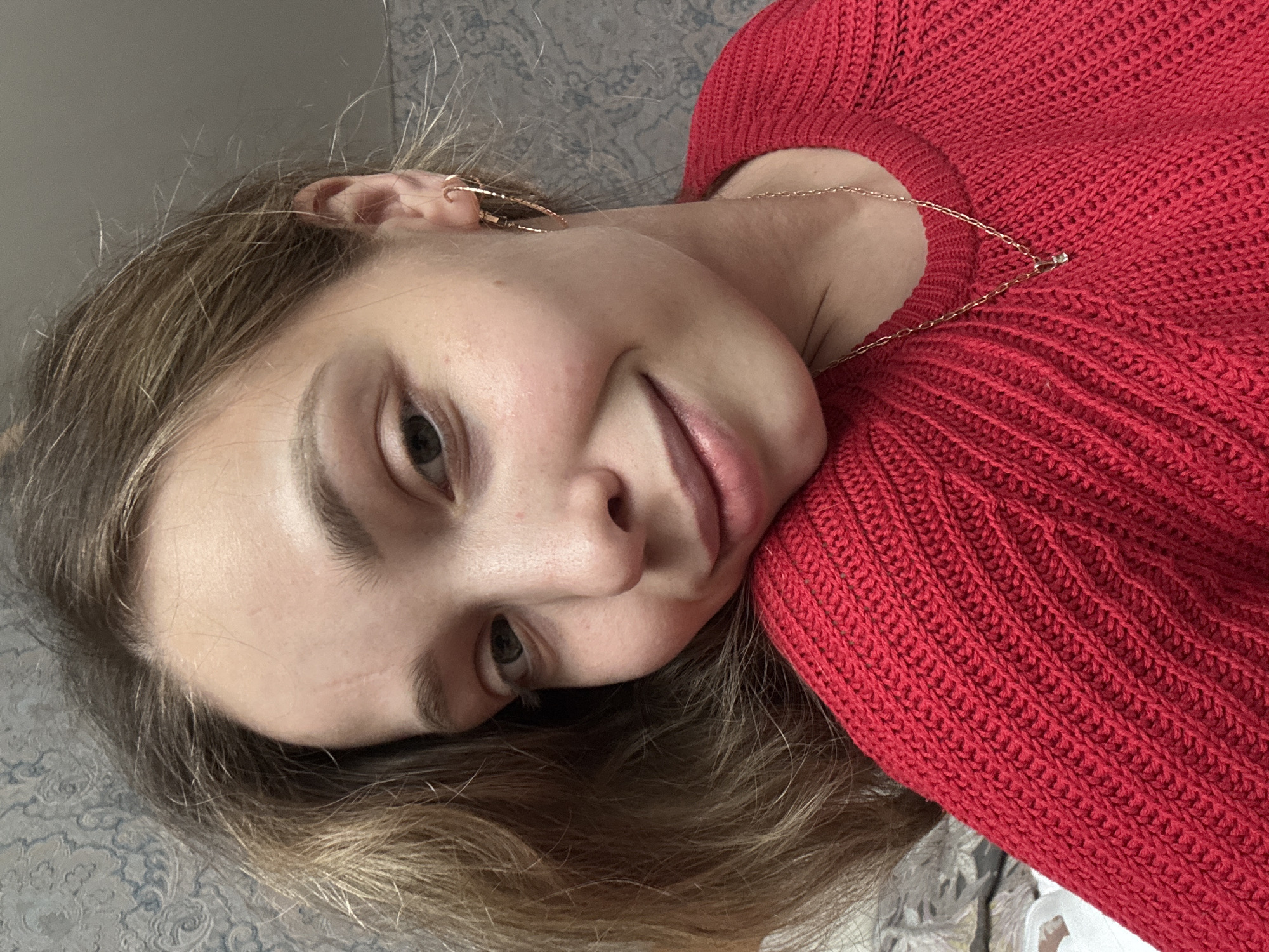 A young woman is lying down, wearing a red knit sweater and gold jewelry, including a necklace and hoop earrings. Her hair is light brown. She has fair skin and a neutral expression. The background is a patterned fabric, possibly a bedspread or couch. There is no information provided to suggest the picture was taken in Прикамский, Russia.