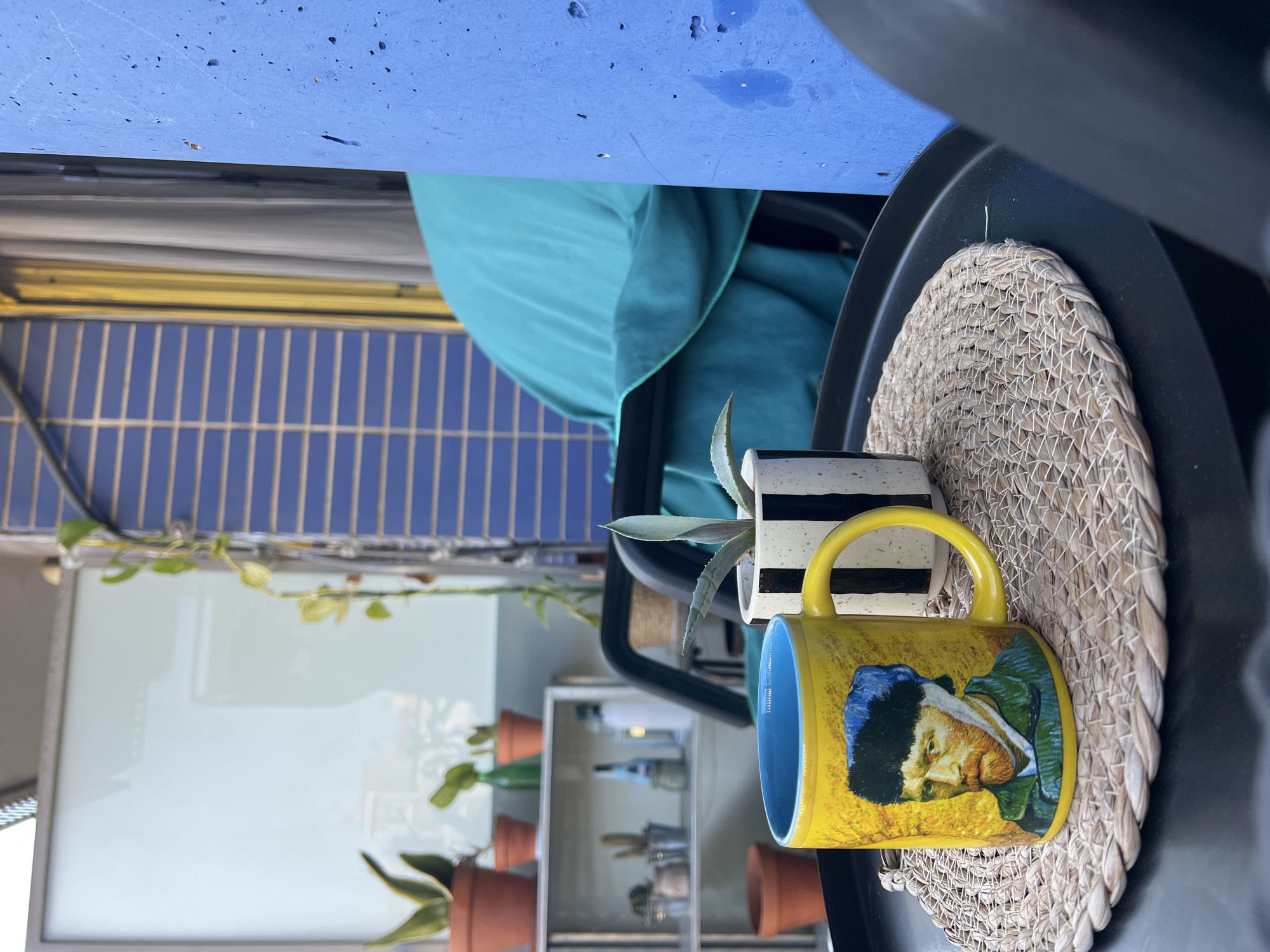 The image captures an outdoor scene in Mollet Del Vallès, Spain. A yellow mug with a blue interior, depicting Vincent Van Gogh, rests on a circular woven placemat atop a black surface. Behind the mug is a small potted plant with a black and white striped pot. In the background, a blue-tiled wall is partially obscured by a light blue fabric draped over something. A shelf with potted plants and various bottles is visible in the background. A trailing vine hangs near a white surface. The scene seems to be on a patio or balcony.