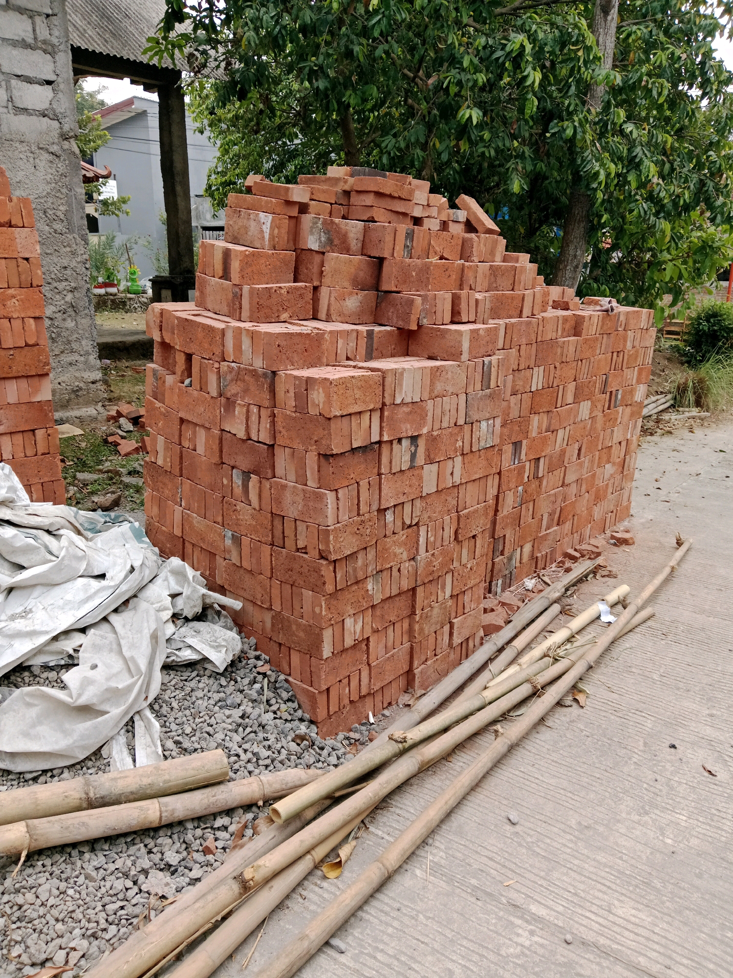The image shows a construction site in Salatiga, Indonesia, with stacks of red bricks in the foreground. The bricks are neatly piled, forming a large rectangular stack. To the left of the brick stack, there are piles of white tarpaulin and gravel. Several bamboo poles are scattered on the concrete ground in front of the bricks. In the background, there is a building with a gray roof, trees with green foliage, and a grey concrete wall. The scene appears to be outdoors on a paved surface, likely a street or yard.