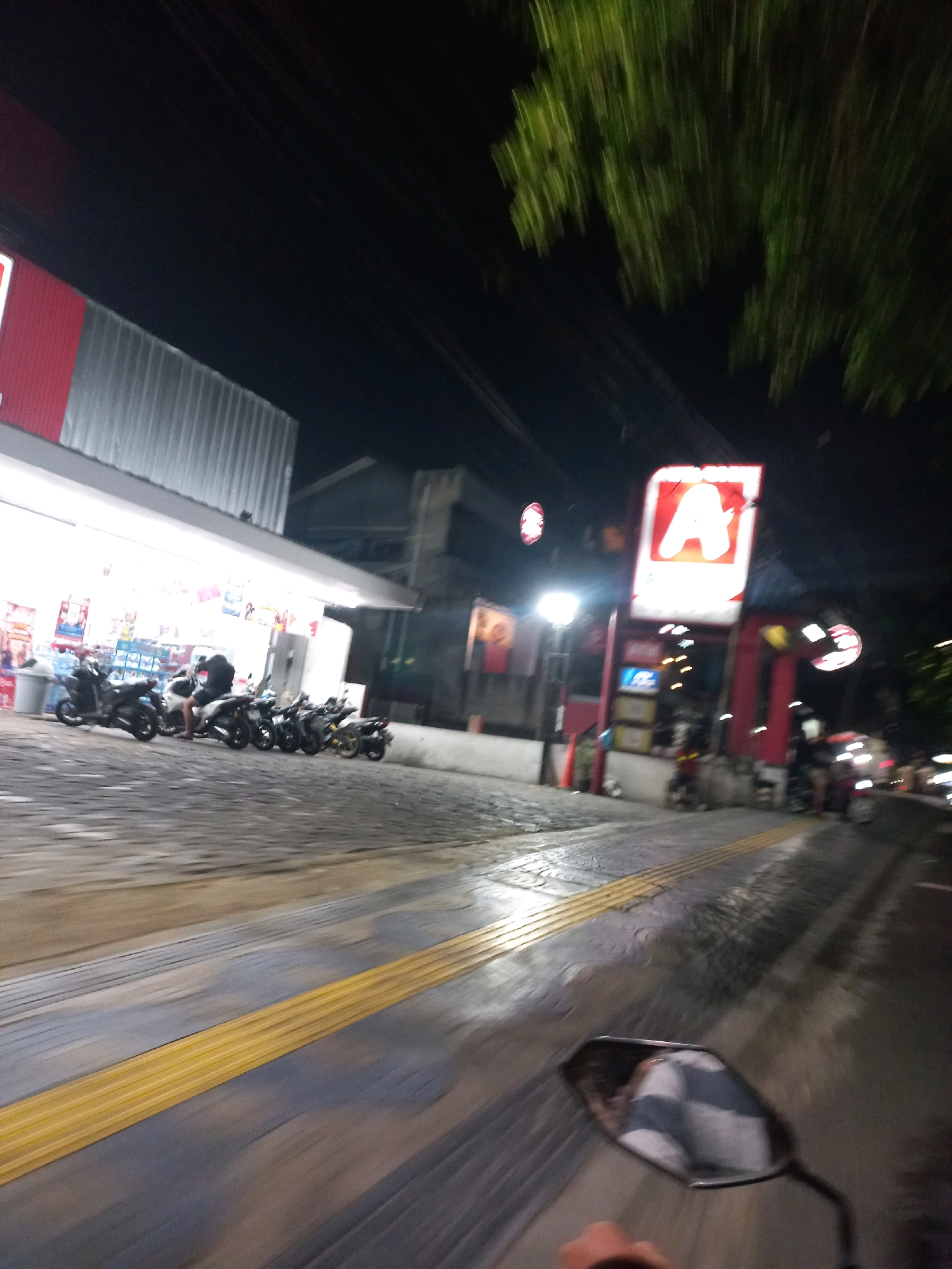 The image captures a nighttime street scene in Depok, Indonesia, likely near a convenience store or small business establishment. In the foreground, a motorcycle's rearview mirror reflects a checkered pattern, suggesting the photographer is on a motorcycle. The street is paved with cobblestones and features a raised yellow tactile paving strip, commonly found on sidewalks or crossings for visually impaired pedestrians.
To the left, a brightly lit store facade is visible, with a corrugated metal awning. Several motorcycles are parked in front of the store, with a few individuals standing or sitting on them. In the background, a prominent red sign with a large white letter "A" is illuminated, indicative of a commercial establishment. The overall environment is dimly lit, characteristic of a street scene at night, with the presence of streetlights and building illumination. Trees with blurred foliage are visible overhead, suggesting an outdoor, urban setting. The motion blur throughout the image indicates that the photograph was taken while in motion, likely from a moving vehicle.