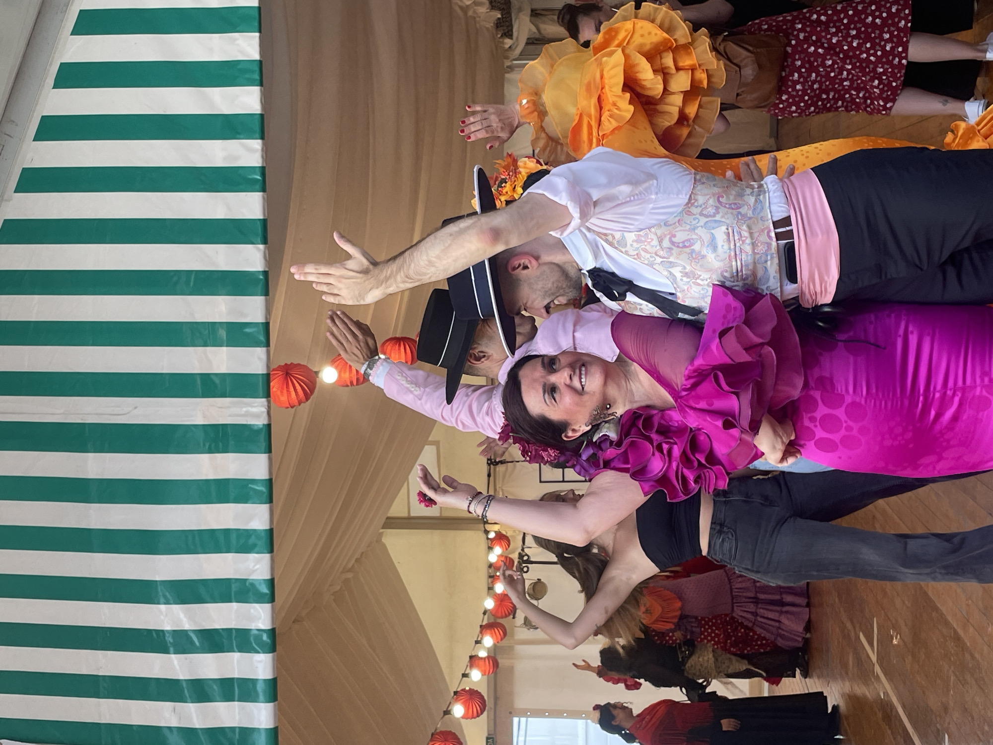 Here is a detailed description of the image:
**Scene Overview:**
The image captures a dynamic scene featuring a group of people dressed in traditional Spanish attire, likely performing or participating in a celebration. The setting appears to be indoors, possibly a tent or hall decorated with festive elements. The location is Sant Adrià de Besòs, Spain.
**People:**
* **Dancers:** Several individuals are visible, primarily positioned to the left and center of the frame. They are wearing flamenco or similar traditional outfits. The costumes showcase vibrant colors like magenta, orange, pink, and black.
* **Men:** One man in the foreground wears a white shirt with a paisley vest, black pants, a pink sash, and a black hat. Another man in the background is wearing a light pink jacket and a similar hat.
* **Women:** At least three women are visible in the foreground. One is wearing a bright magenta dress with ruffled sleeves and neckline. Another is wearing dark jeans and a black top with an orange and purple skirt. The third woman is wearing an orange dress with large floral embellishments.
**Objects and Environment:**
* **Decoration:** The background includes a beige fabric backdrop, possibly the tent walls. Red paper lanterns are suspended, adding to the festive atmosphere. A green and white striped structure, likely part of the tent's support or decoration, is visible on the left.
* **Attire:** The costumes include elements such as ruffled sleeves, floral headpieces, vests, hats, and sashes. These are characteristic of Spanish traditional dress.
* **Flooring:** The floor appears to be made of wood.
**Actions and Interactions:**
* **Performance:** The people are striking poses, indicating a dance or performance.
* **Expressions:** The people appear joyful and energetic.
* **Group Dynamics:** The photo suggests a communal activity or celebration, with the individuals engaging in a shared performance.
