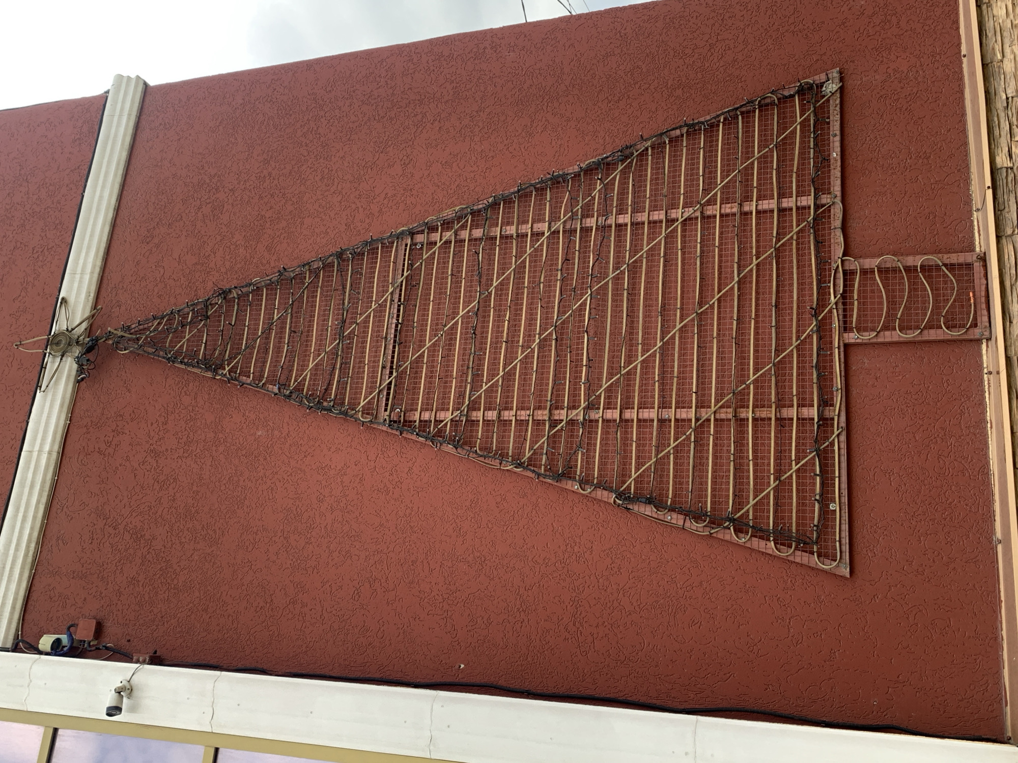 The image shows a section of a reddish-brown exterior wall, textured with a stucco-like finish. Affixed to the wall is a triangular metal frame filled with vertical, tan-colored tubes or rods. Black wiring, possibly lights, are wrapped around the perimeter of the triangle. To the right of the triangle, another rectangular metal frame is visible. Below, a security camera is mounted near the bottom of the wall. A narrow white molding runs vertically up the left side of the wall. The location is Stavropol, Russia.