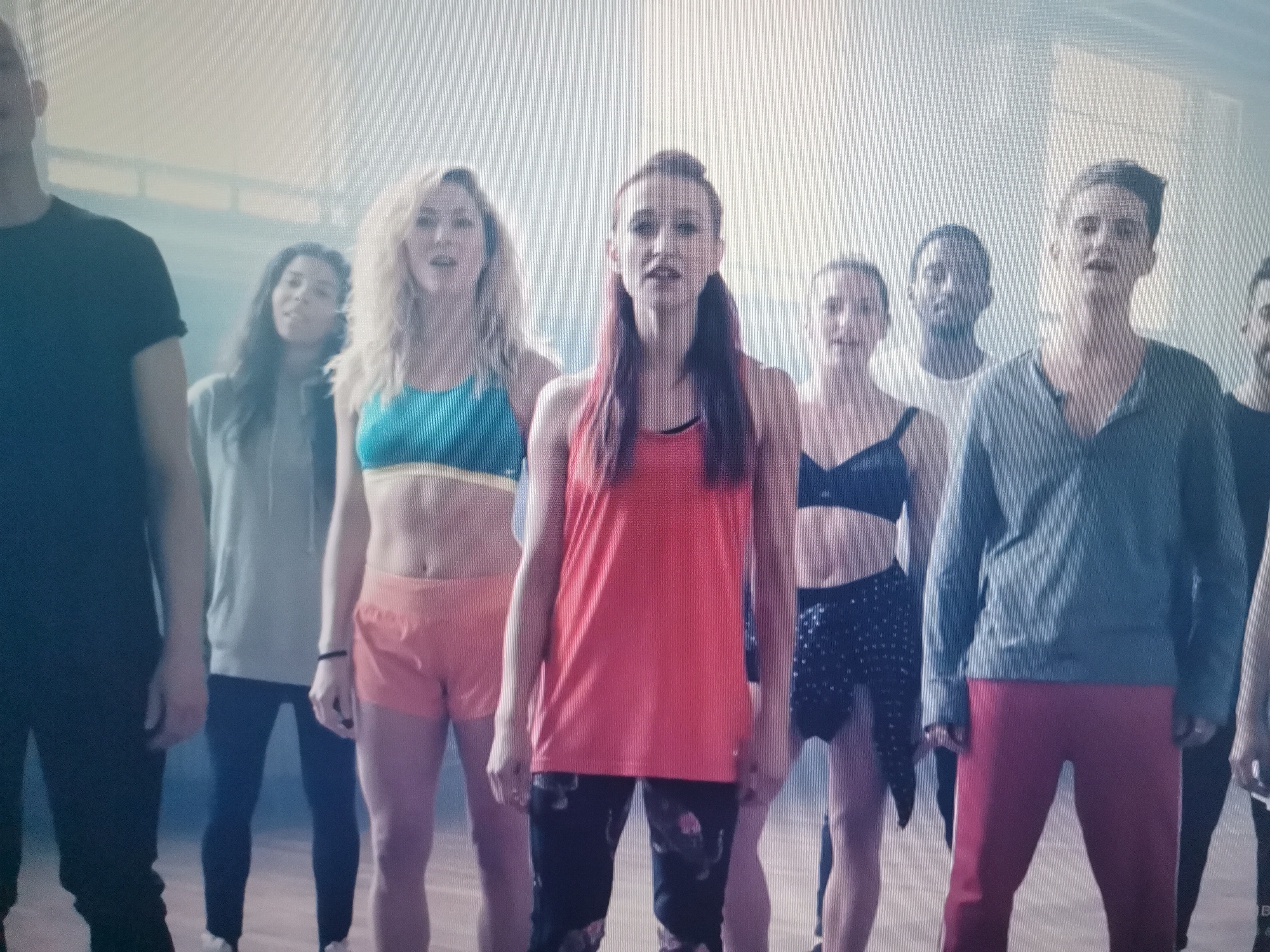 The image depicts a group of ten individuals, appearing to be in their late teens to mid-twenties, standing in a loosely formed line or cluster. They are inside what looks like a large, sparsely furnished room, possibly a dance studio or gymnasium. The background is largely indistinct due to a hazy, bright light, but suggests industrial-style architecture with large windows or openings. The flooring appears to be wood.
The individuals are dressed in athletic or casual wear, with some wearing tank tops and shorts or leggings, and others in t-shirts and long pants. Their postures suggest they are either posing or engaged in a group activity. Some of them are looking directly at the camera.
There is no visible indication of the city of Belaazyorsk, Belarus in the image.