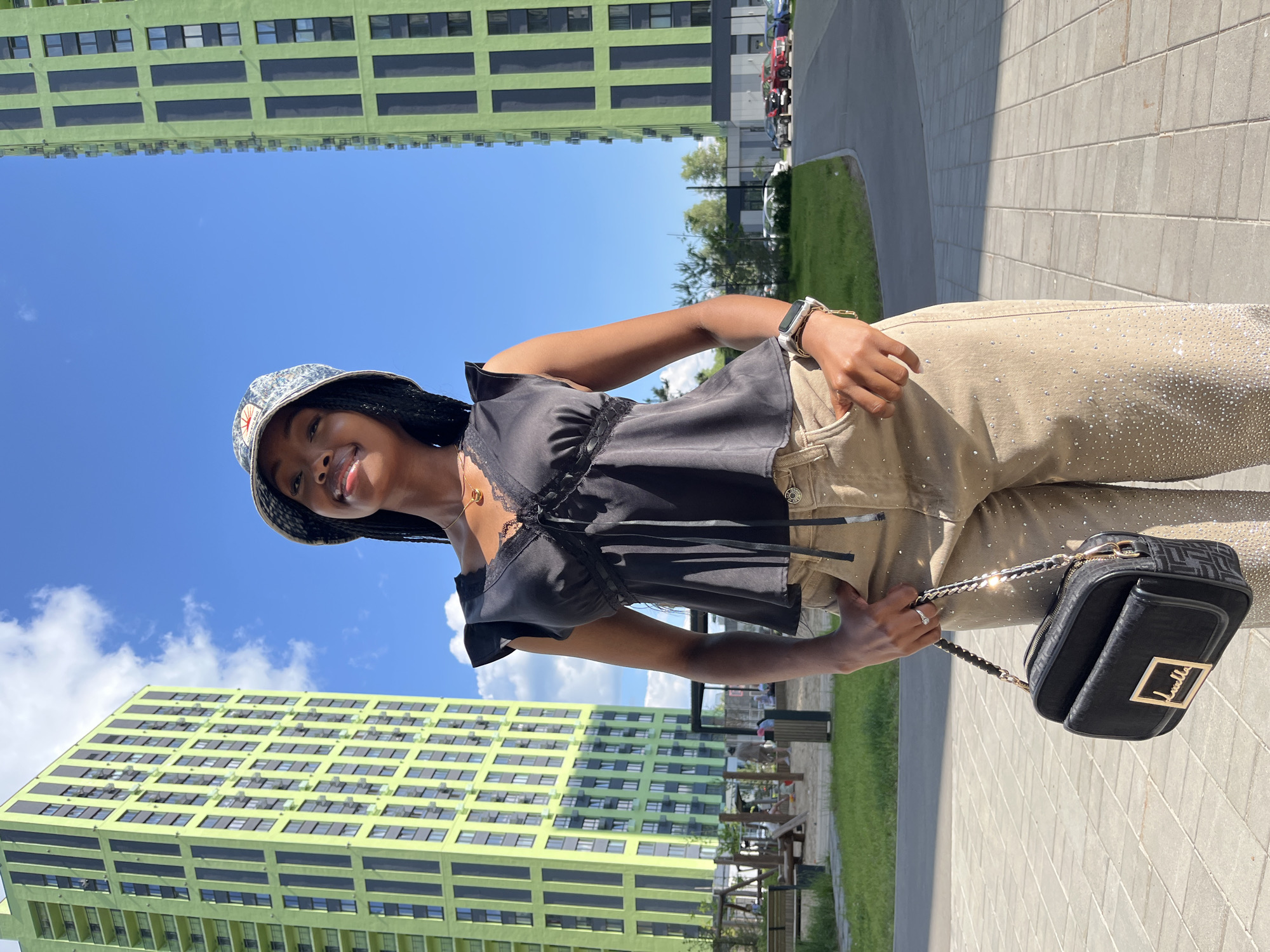 A Black woman stands outdoors, facing the camera, in front of two tall, lime green buildings with dark vertical accents. The sky above is blue with scattered white clouds. She is wearing a black ruffled sleeveless top with lace trim and a bucket hat. Her pants are tan and appear to be decorated with rhinestones. She holds a black purse with a gold chain strap in her left hand. Her right hand is in her pocket. The ground is paved with gray square tiles. In the background, to the left, a playground with wooden structures is visible. To the right, there is a road with parked cars. The scene is likely in a modern urban or residential area in Kazan, Russia.