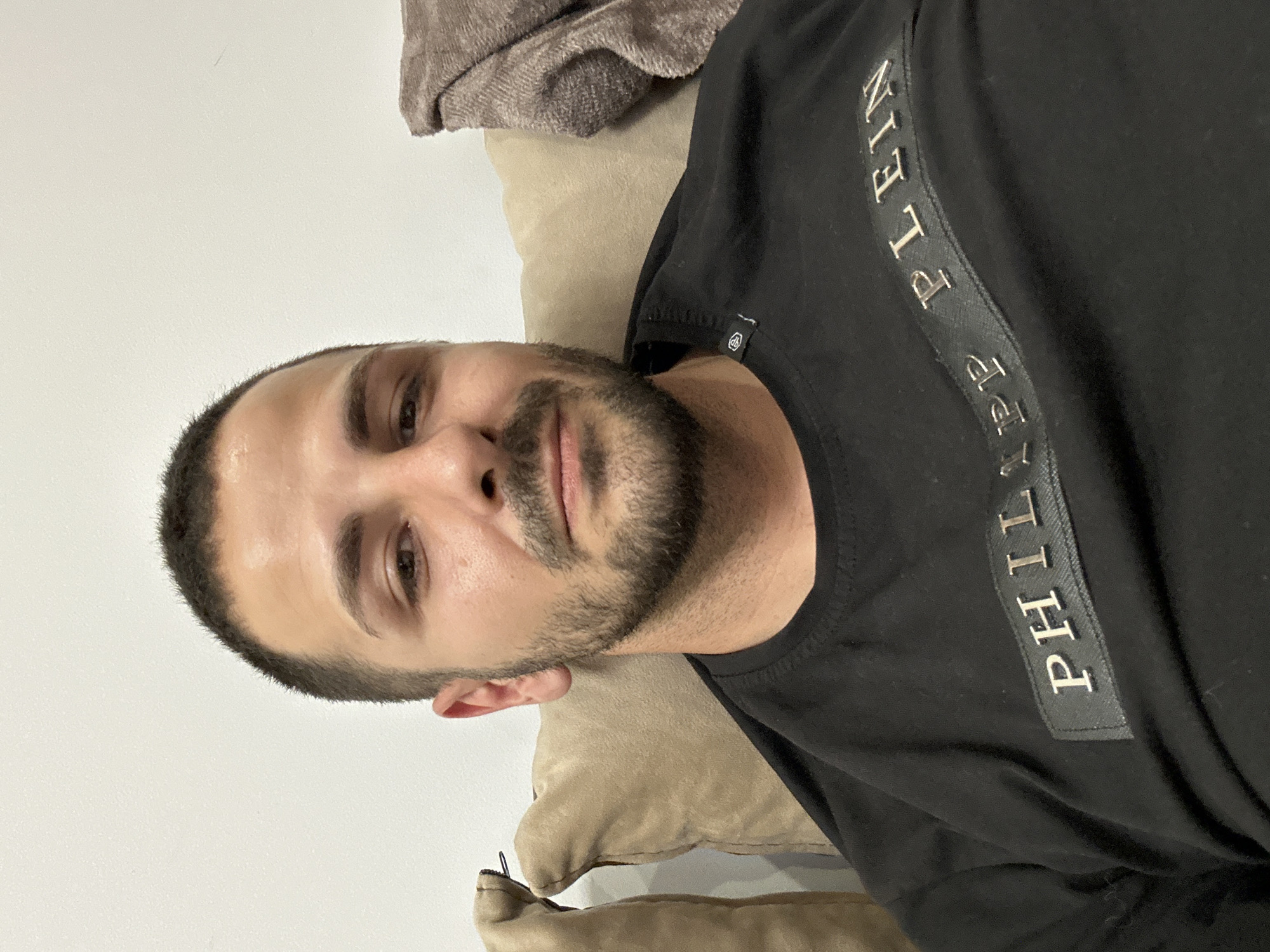 A medium shot depicts a man indoors, reclining against beige cushions. He has a closely-cropped haircut and a short beard. The man is wearing a black t-shirt with the words "PHILIPP PLEIN" printed vertically on the front in gold letters. The location appears to be a private residence, city Unknown, Unknown.