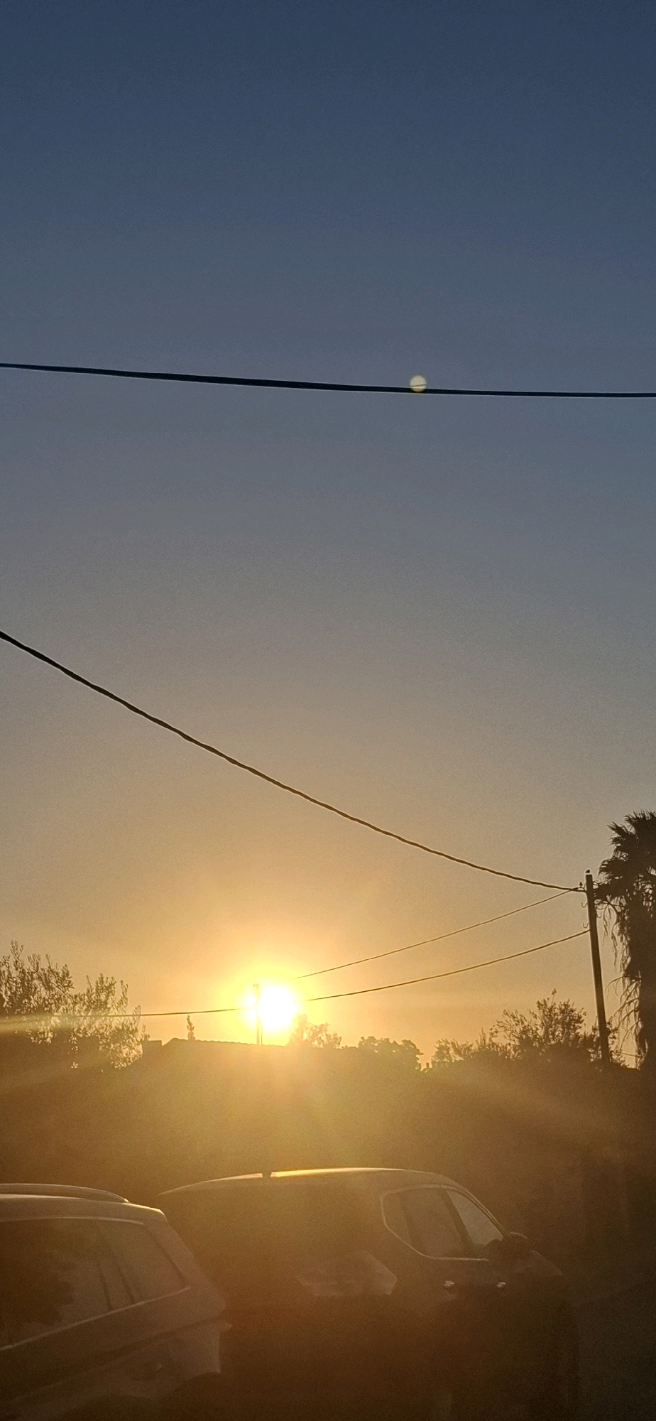 The image shows a sunset in Yerocham, Israel. The sun is low in the sky, casting a warm, orange glow over the scene. Power lines stretch across the frame, silhouetted against the vibrant sky. In the foreground, two vehicles are parked, their shapes softened by the atmospheric light. Trees and buildings are visible in the distance, partially obscured by the haze and lens flare created by the setting sun. The overall impression is one of a peaceful evening in a quiet desert town.