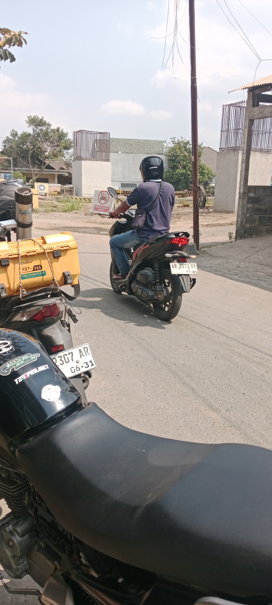 The image, taken on a sunny day in Purwomartani, Indonesia, captures a street scene with several motorcycles. In the foreground, the black seat and engine details of a motorcycle are visible. Behind it, another motorcycle is parked, featuring a yellow storage box labeled "tumtec FST-16S CORE FUSION SPLITTER". A third motorcycle, ridden by a person wearing a blue shirt, dark helmet, and jeans, is in motion, its license plate reading "AB 6573 UX 07.29". The rider is seen from the back, with a small bag slung over their shoulder. The background reveals construction elements, including concrete pillars and rebar structures, along with buildings and trees under a bright sky with scattered clouds. A traffic sign with a red circle and diagonal line indicating a prohibition is also visible.