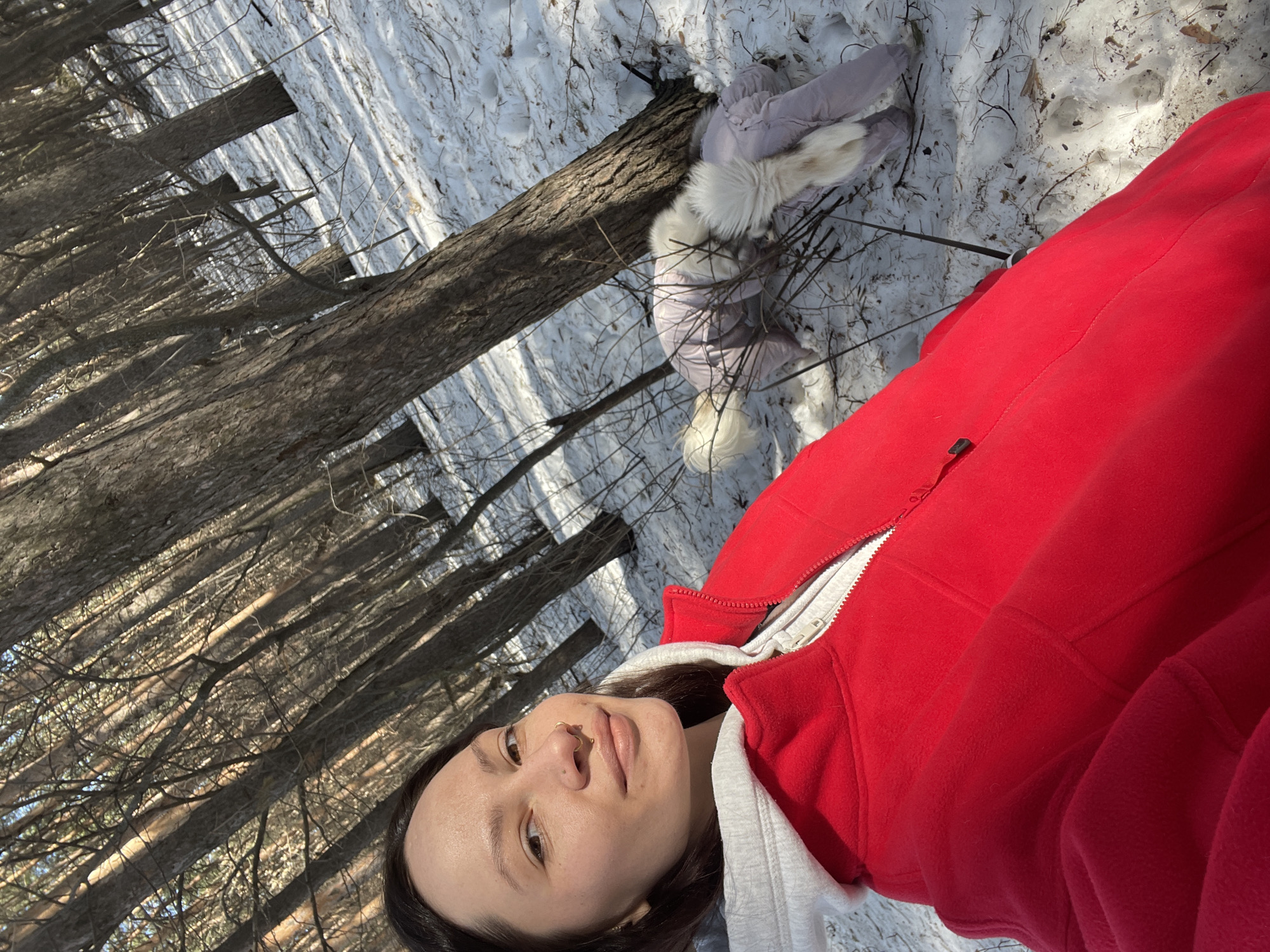 A woman in a red jacket takes a selfie in a snowy forest. A light-colored jacket with fur trim is near a fallen tree. The location is identified as a forest in Izhevsk, Russia.