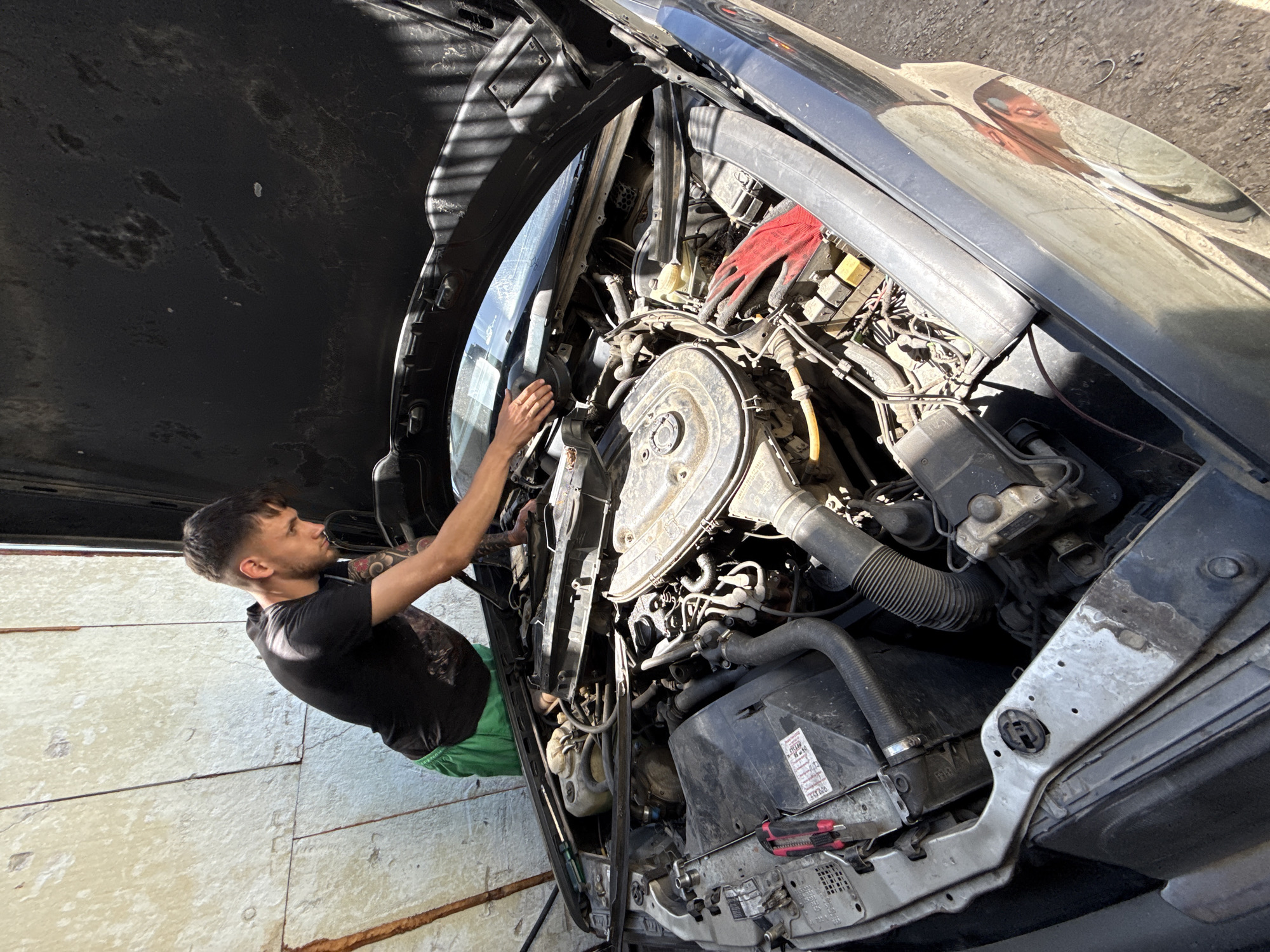 A man is working on a car engine with the hood open. He is wearing a black t-shirt and green shorts. The car is parked in a garage or workshop with light-colored stone or concrete walls. The reflection in the car shows what seems to be a person standing nearby. The environment looks like an urban area, which may be in Shumen, Bulgaria. The engine is old and greasy and cluttered with various components. There are red gloves laying on the engine.