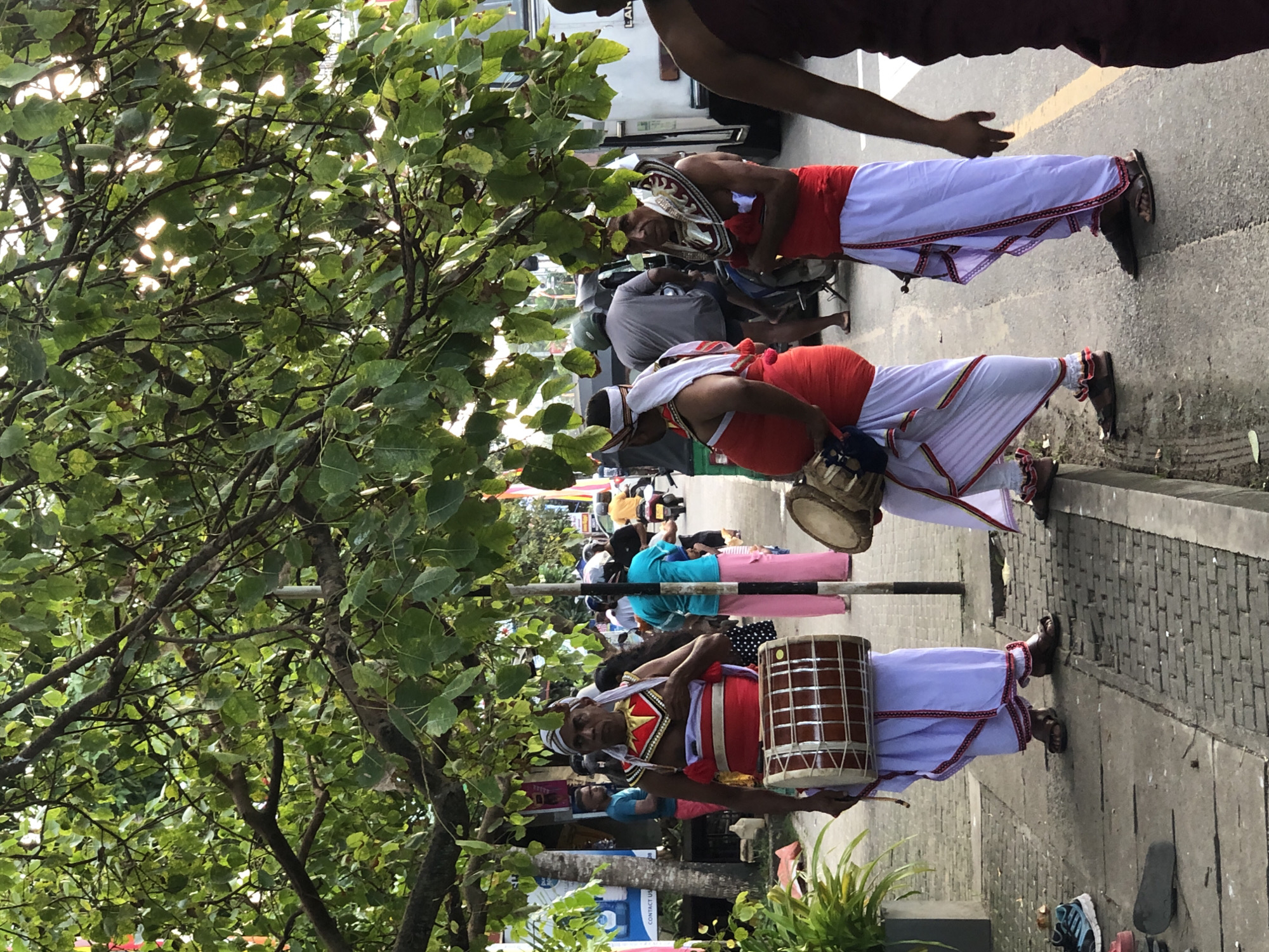 The image captures a street scene in Habaraduwa, Sri Lanka, featuring several individuals participating in what appears to be a cultural performance or procession. Two men in traditional attire, red vests and white sarongs with decorative borders, are prominently featured. One is playing a drum. Another man, also in traditional clothing, is visible in the background with a drum. A person in the upper portion of the image wears a dark red garment.
The setting is a paved street with some greenery from a tree in the foreground. The background shows a street with vehicles, including motorcycles, and other people. The lighting suggests it might be daytime, possibly late morning or afternoon, with some dappled sunlight filtering through the leaves of the tree. The overall mood seems lively, possibly a festive occasion. The image is oriented vertically, with the street running from the bottom to the top of the frame, and the tree on the left. Some of the clothing has intricate patterns and colorful accents. There are no other significant visible text elements.