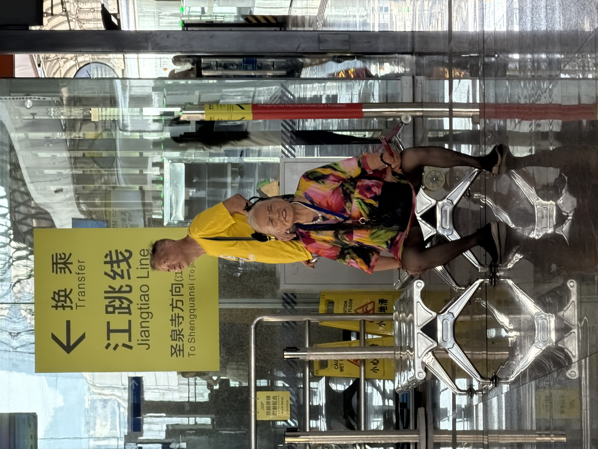 The image depicts an indoor scene, likely a transportation hub, with a shiny, reflective floor. In the foreground, an elderly woman is seated on a metallic bench. She is wearing a brightly colored, floral-patterned dress, black pantyhose, and black shoes. She has a necklace with a pendant and is holding a small red object in her right hand. Behind her, standing to her left, is a man wearing a yellow t-shirt. To the left of the man, a yellow sign with black Chinese characters and English text "Transfer" and "Jiangtiao Line" is visible. The sign also indicates "To Shengquansi (To Jiatiao)". Further to the left, there are metal barriers and yellow caution signs. The environment appears to be a modern interior with glass walls and metallic structures. The overall impression is that of a waiting area in a transit station in Tiaodeng, China, given the signage.
