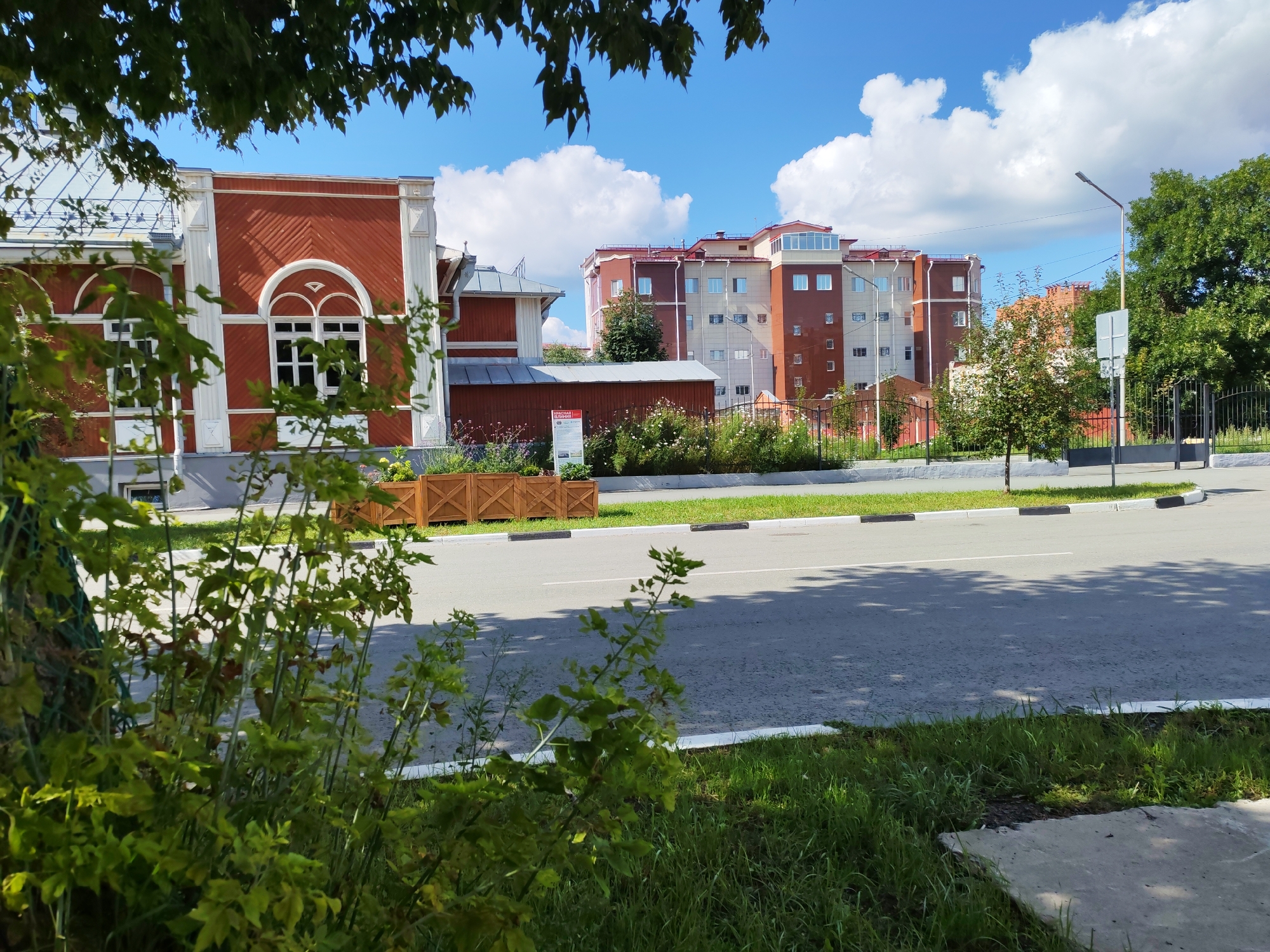 The image shows a street scene in an urban environment under a bright blue sky with scattered white clouds. In the foreground, lush green foliage partially obstructs the view of a road and sidewalk. The road has a white line marking its edge and a white center line. Shadows of trees are cast across the asphalt.
Along the side of the road, there is a strip of grass and a concrete sidewalk. Further back, a dark metal fence runs parallel to the road. Beyond the fence, a building with red and white architectural details, featuring arched windows, is visible. Next to it, a more modern, multi-story building with a reddish-brown and beige facade stands tall. Trees are interspersed between the buildings and along the fence line. A lamppost is visible to the right of the multi-story building. Wooden planters are placed along the sidewalk in front of the red and white building, filled with greenery and some flowers. A sign is attached to one of the planters.