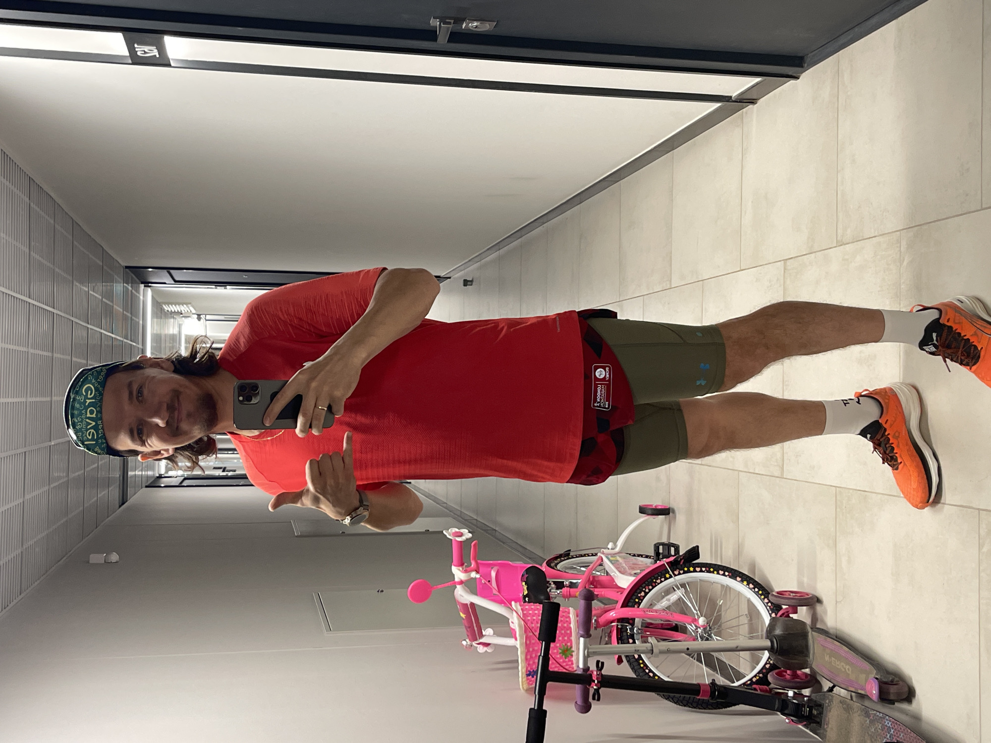 A man wearing a red shirt, olive-green shorts, orange running shoes, a green patterned head covering, and a watch on his left wrist is taking a selfie in a tiled hallway. He is holding a smartphone in his right hand and gesturing with his left hand. Behind him are a pink bicycle and a scooter. The hallway has gray walls and ceiling with dark trim. A sign with Russian characters is visible at the end of the hall. The image appears to be taken indoors, likely a residential or commercial building in Izhevsk, Russia.