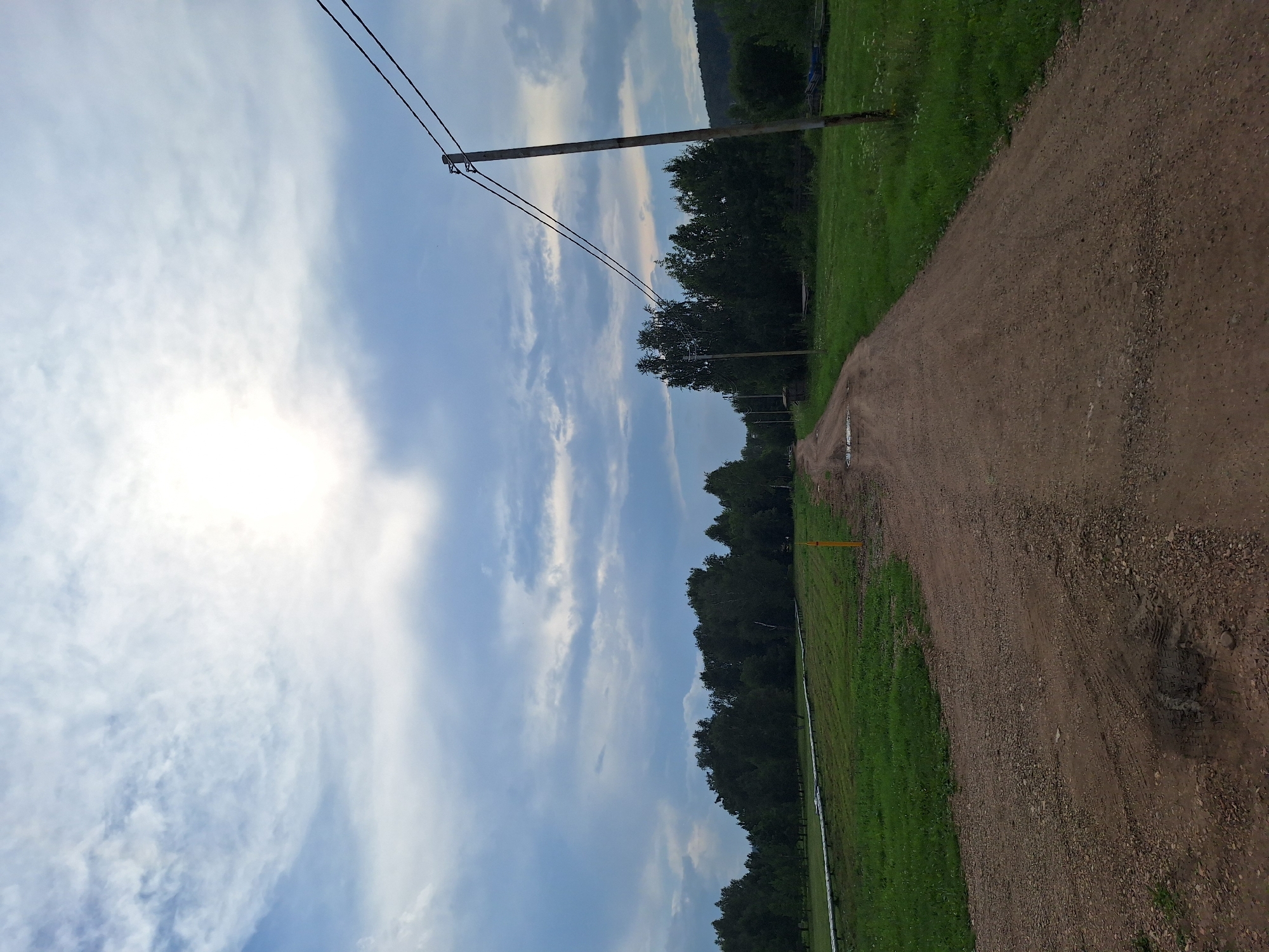The image shows a dirt road winding through a rural landscape. On the left side of the road, there is a green grassy field with a long white pipe running through it. A yellow marker post is visible in the field. A line of trees forms a dark green border on the horizon behind the field. On the right side of the road, a wooden utility pole with two wires stretches across the frame. Beyond the pole, more trees and a grassy area are visible. The sky is partly cloudy with the sun breaking through, casting a bright light. The overall impression is one of a rural, undeveloped area, possibly in the city of Atamanovka, Russia.