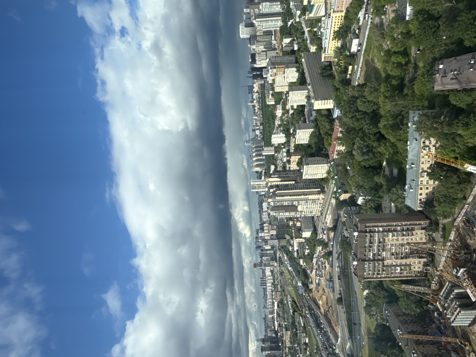 The image depicts an aerial view of a city, likely Khimki, Russia, under a dramatic sky. A distinct line of clouds separates a clear blue sky from an area of dark, ominous storm clouds. Below, the city unfolds with its dense urban environment of buildings, roads, and green spaces. The infrastructure details include a network of highways and railways, suggesting a major transport hub. Buildings range in size and style, from apartment blocks to taller structures, potentially office buildings. Construction is evident in the lower right corner, where cranes and partially built structures stand. There are no people immediately visible in the overview.