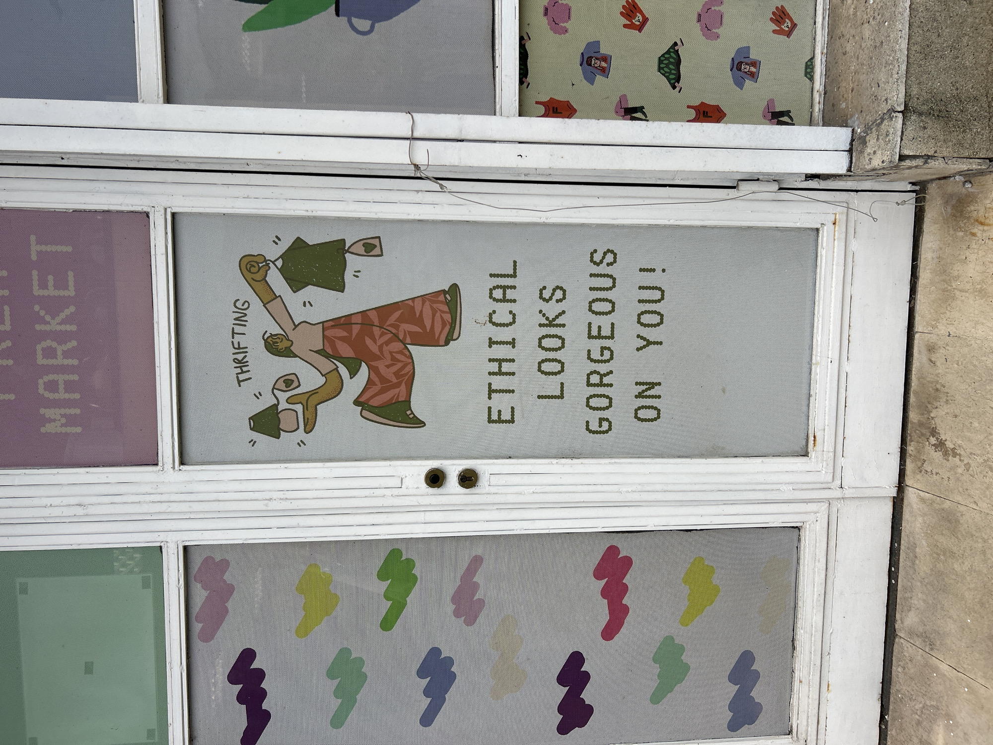 A street-level, close-up photograph shows a white-framed door or window with multiple panes. The panes contain various images and text. From left to right, top to bottom, the panes feature: partial text ending in "MARKET"; an illustration of a person holding up clothes with the text "THRIFTING ETHICAL LOOKS GORGEOUS ON YOU!"; a pattern of clothing images; a plain green square; a pattern of colorful lightning bolt shapes. The door appears slightly aged with worn paint. The photograph appears to have been taken during the day. The location, Plovdiv, Bulgaria, is unknown from the image.