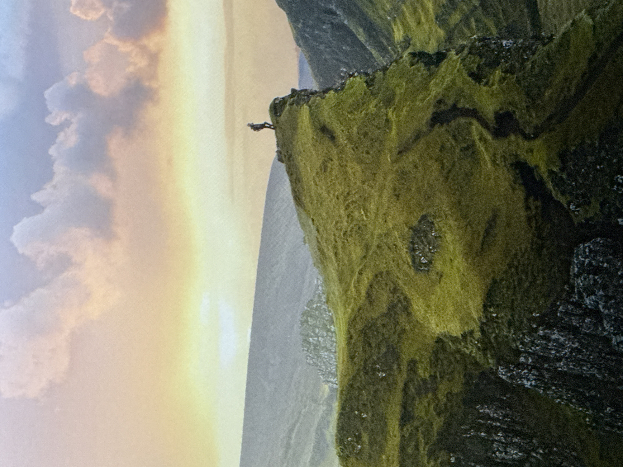 The image depicts a lone hiker standing on the edge of a grassy cliff, overlooking a vast, mountainous landscape. The sky above is a gradient of soft, pastel colors, transitioning from pale yellow near the horizon to a hazy blue at the top, with wispy clouds. The terrain is rugged, with rolling hills covered in vibrant green grass, interspersed with darker patches and rocky outcrops. A winding path is visible on the side of the cliff. The scene evokes a sense of solitude and adventure. This image is not located in the city of Saratov, Russia; it is a natural landscape scene.