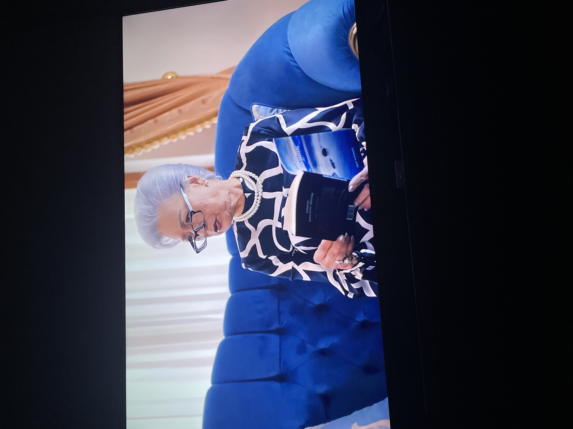 An elderly woman with gray hair styled up, wearing glasses and a pearl necklace, is sitting on a plush royal blue sofa. She is wearing a black and white patterned dress and is holding an open book with a picture of a beach on the cover. The background shows light-colored drapes and a decorative golden trim. The scene appears to be indoors, possibly in a living room or a studio. There is no visible indication of the location being in Tallinn, Estonia.
