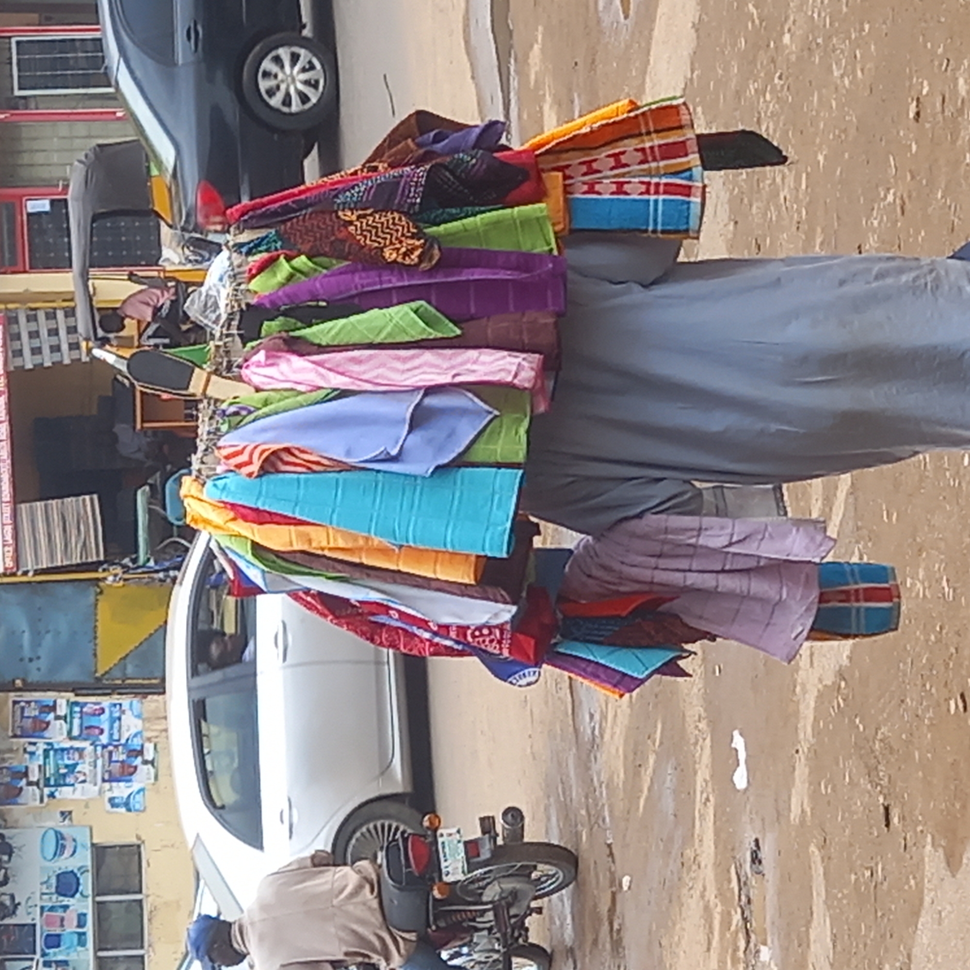 In Dadi Riba, Nigeria, a person is carrying several stacks of folded, colorful towels on their head and shoulders, standing on a street. To their left is a white car and a motorcycle. Behind them is a dark car, with shops visible in the background. The ground appears to be pavement.