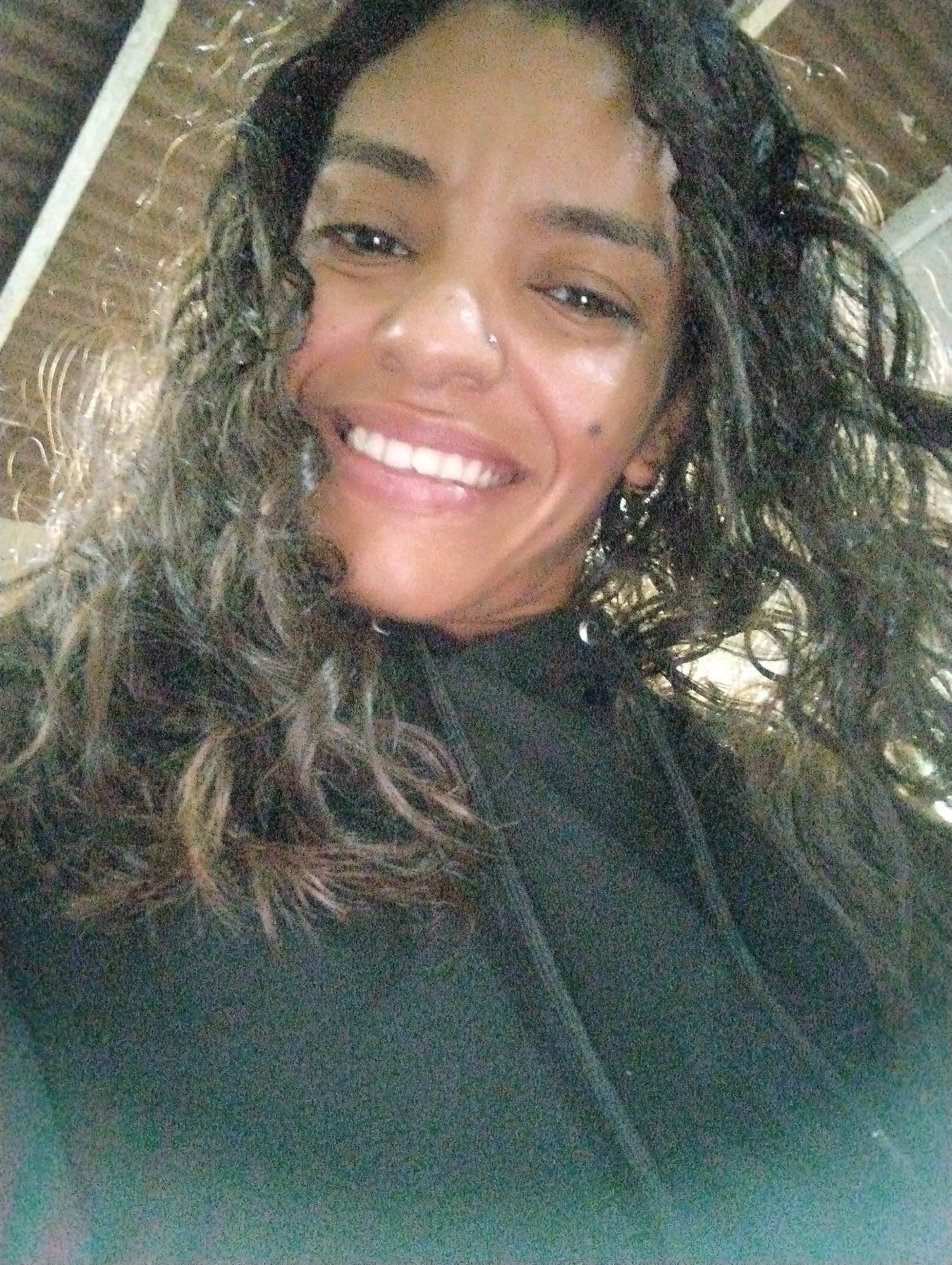 A woman is smiling and looking directly at the camera. She has curly hair and is wearing a black hoodie. The background is not clear, but there are some structural elements suggesting an indoor or covered space. The location is El Valle del Espíritu Santo, Venezuela, but the image does not offer any details to confirm this.