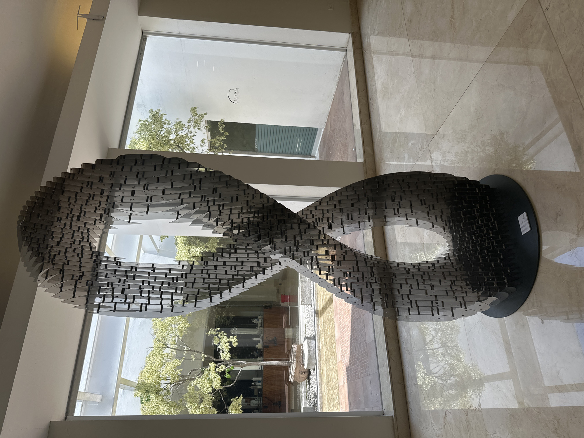 The image showcases an indoor architectural space, likely a modern lobby or gallery, featuring a prominent sculpture. At the center is a large, metallic sculpture resembling an infinity symbol or a Mobius strip. It is intricately constructed from numerous small, interconnected dark grey pieces, creating a textured, layered surface, and rests on a flat, black circular base. The indoor space features light-colored walls and a highly reflective, light-toned tiled floor, possibly marble, which mirrors both the sculpture and the exterior view.
Large, clear windows in the background provide a bright view of an outdoor courtyard or garden. Through these windows, trees with vibrant green and variegated foliage are visible, along with sections of white or light-colored building walls. The ground outside appears to be a mix of paved areas, including brickwork. The bright natural light streaming in suggests it is daytime, with clear weather conditions.
No people are present in the scene, conveying a sense of tranquility and artistic contemplation. A reflection of the words "LA VISTA" is faintly visible on a white wall through one of the windows, possibly indicating a business or property name. Unreadable text is also visible on a small label on the sculpture's base. The location is identified as San Bernardino Tlaxcalancingo, Mexico.