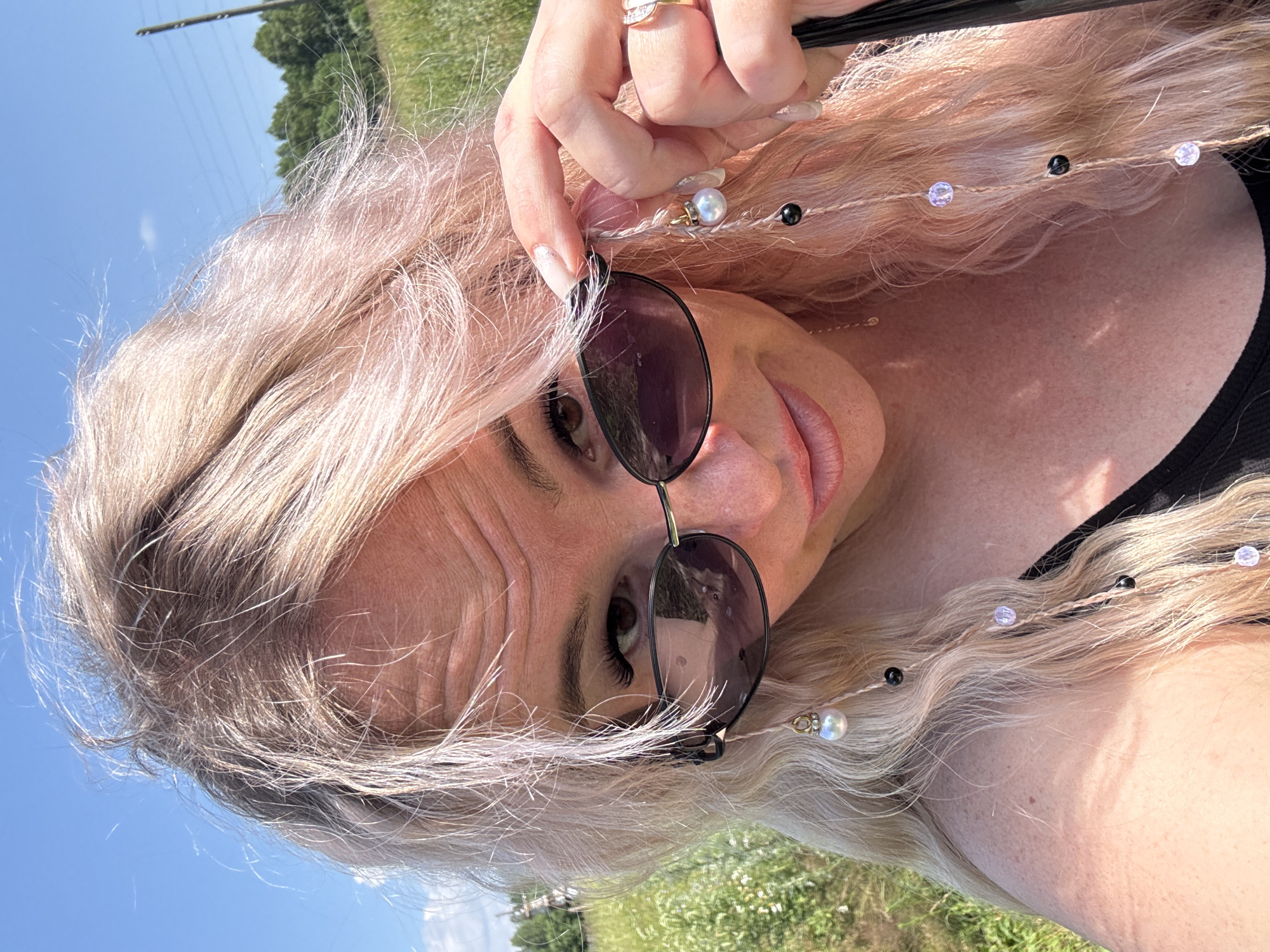 In a candid, close-up selfie, a fair-skinned woman with long, wavy, light pink hair is positioned outdoors. She wears dark, oval-shaped sunglasses and has on a black tank top. Her right hand is raised, with her index finger and thumb gently adjusting strands of her hair that are intertwined with thin strings adorned with beads, including pearls, black beads, and clear iridescent beads. Her fingernails are painted with a light, shimmering polish. A ring with a light-colored stone is visible on her right ring finger. Her gaze is directed towards the camera, and her lips are slightly parted. The background consists of a blurred, sunny outdoor scene with green grass, foliage, and a bright blue sky with a few wispy clouds. The overall impression is one of a casual, sunny day. The location is identified as Pozdyshki, Russia.