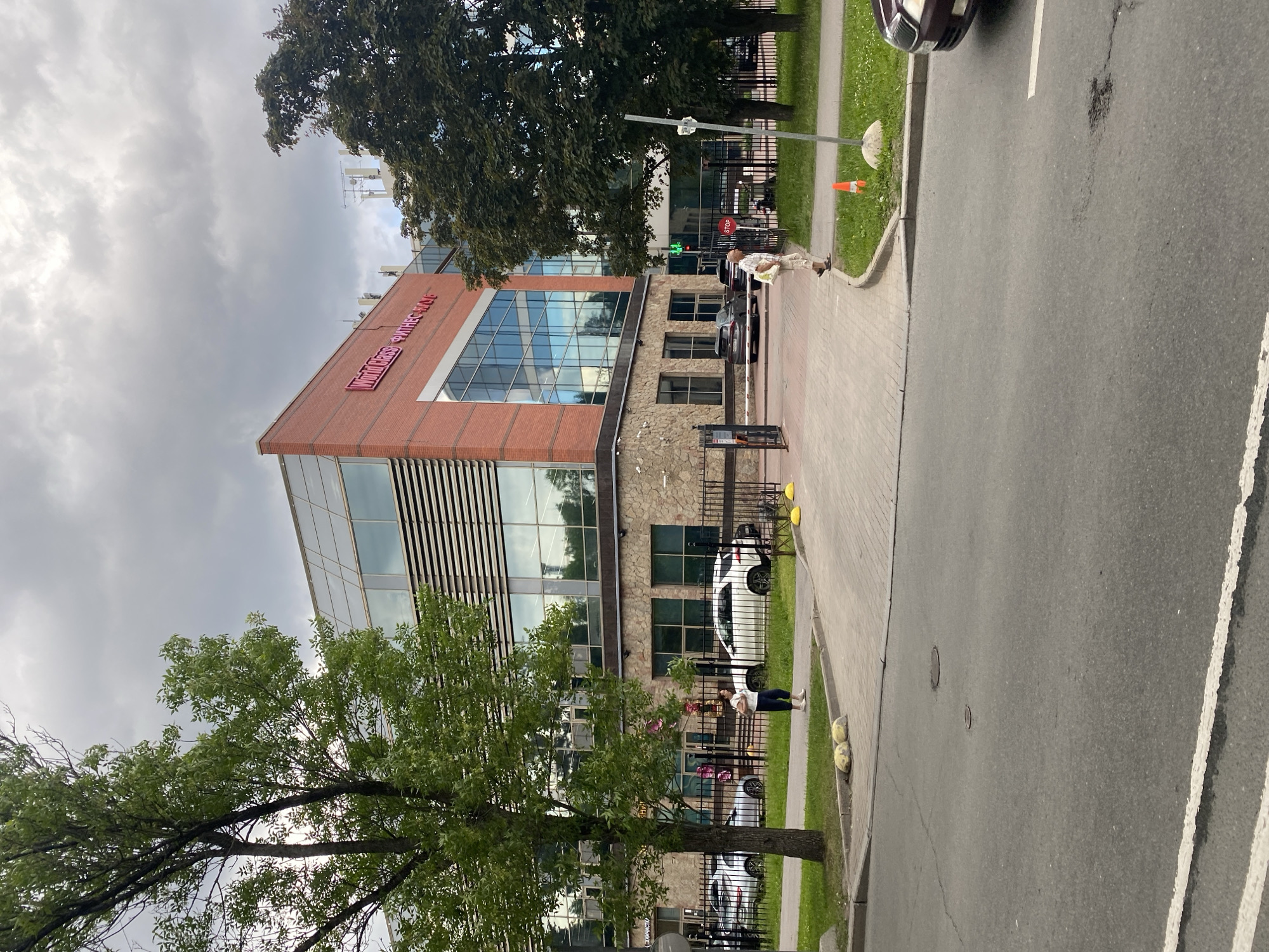 The image depicts the exterior of a modern brick and glass building, identified by a red sign reading "World Class ФИТНЕС КЛУБ" (World Class Fitness Club). The building features large glass windows and a stone facade on the lower level. A tree with green foliage stands in the foreground on the left, partially obscuring the building. A paved walkway leads to the entrance of the building, bordered by a metal fence. Two individuals are visible on the walkway; one woman in a white top and dark pants is walking away from the camera, and an older man in a light-colored shirt and pants is walking towards the camera carrying a bag. Several cars are parked or driving in the background, including a white SUV and a dark sedan. The scene is set on a cloudy day, with a street and sidewalk in the foreground. The location is Saint Petersburg, Russia.