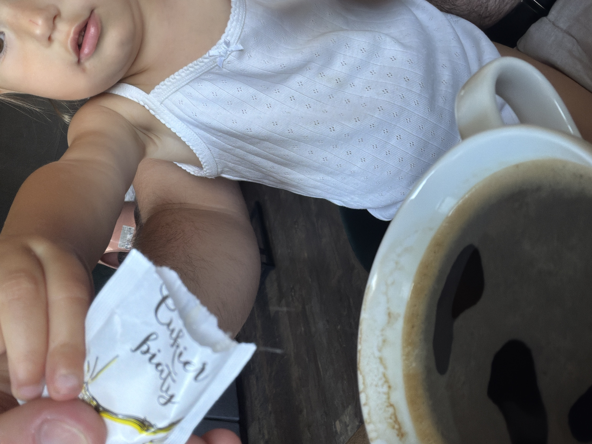 An adult hand, visible on the left, holds a small, torn white packet labeled "Cukier Biały" (White Sugar) with a yellow graphic. White granular contents are actively falling from the packet towards a white ceramic cup, which is partially visible at the bottom of the frame. The cup contains dark liquid, consistent with coffee, exhibiting some surface foam. A child, partially depicted on the right, observes the action. The child has fair skin, visible facial features including the mouth, nose, and a portion of the right eye, and is wearing a white, textured camisole with a small bow near the right shoulder strap. A portion of the child's arm is also visible. The objects rest on a dark, flat surface. The scene takes place indoors, in the city of Sosnowiec, Poland.