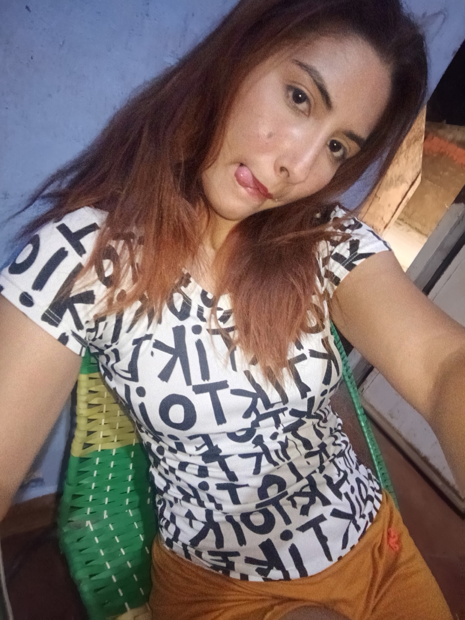 The image shows a woman sitting in a green plastic chair. She is wearing a white t-shirt with black lettering and orange shorts. She is sticking her tongue out. The background is a blue wall on one side and a white wall on the other. The location is Juan Griego, Venezuela.