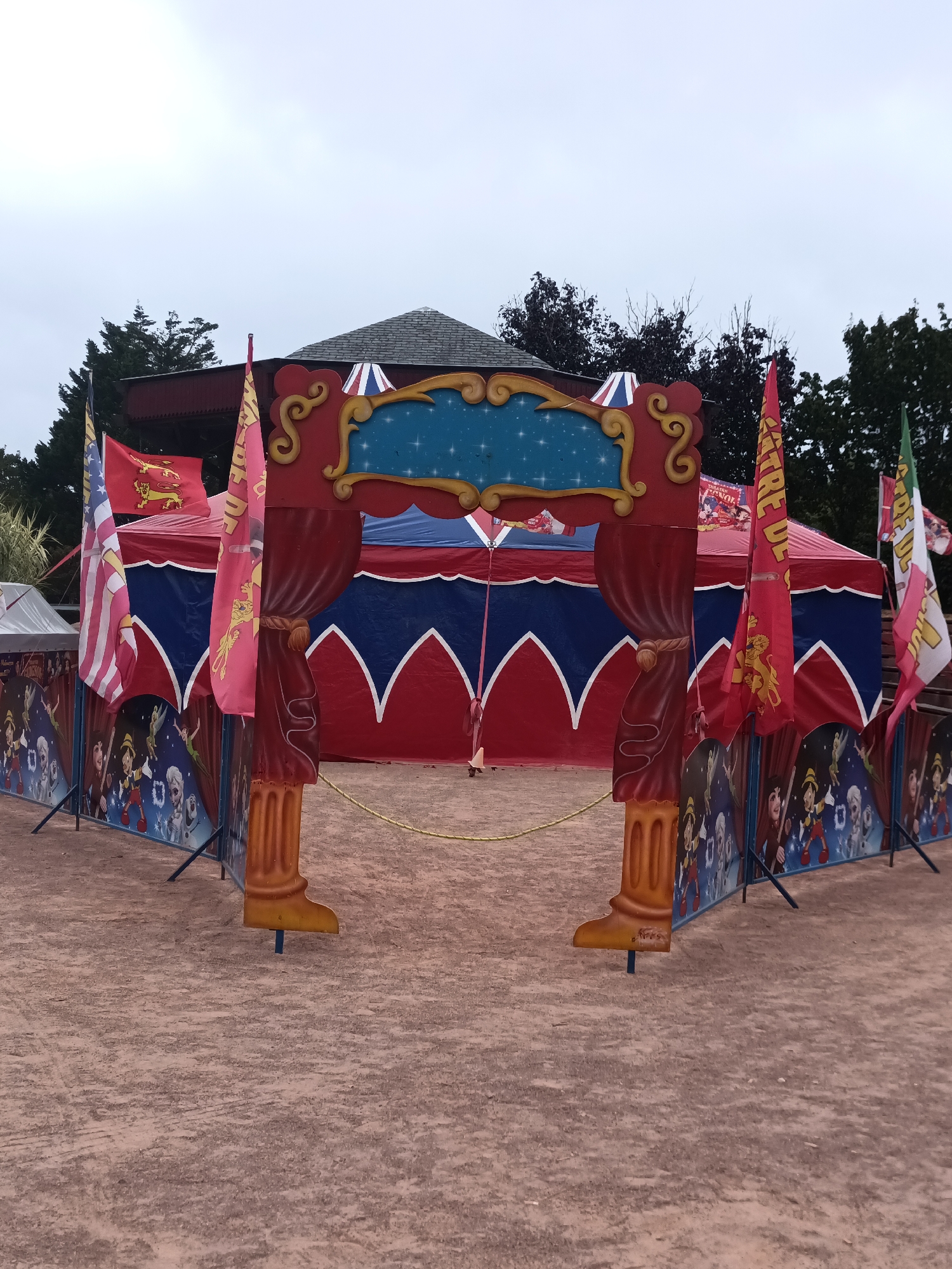 The image shows an outdoor view of a small circus tent with a colorful entrance archway. The tent itself is red and blue with white scalloped trim. Flags are flying from poles around the entrance. The entrance arch is ornate, with red and gold detailing, and features a blue, star-filled background area. Below the arch and extending around the front of the tent is a decorative barrier with images of cartoon characters, possibly from a children's show like "The 101 Dalmatians" and "Pinocchio". The ground is made of reddish-brown dirt. Trees are visible in the background, and the sky is overcast. The overall impression is a festive, albeit somewhat weathered, setup for a small entertainment event. This scene is located in Houlgate, France, as indicated by the flags with the coat of arms of Normandy, and the general architecture of the tent.