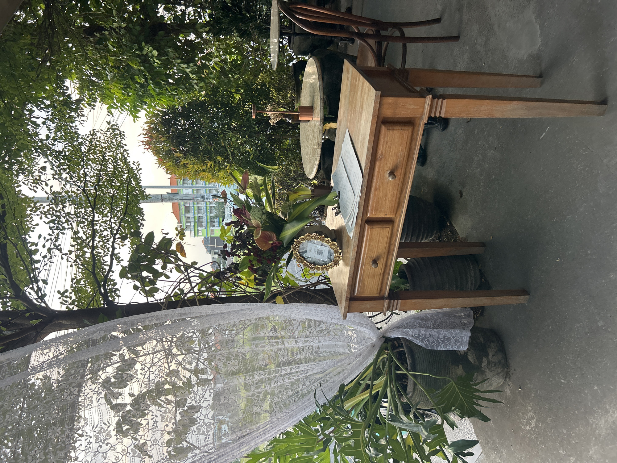 The image depicts an outdoor seating area at a cafe in North Kuta, Indonesia. A wooden table with two drawers is centrally placed, adorned with a bouquet of flowers in a vase and a small, ornate gold-framed picture. A stack of papers is clipped together on the table. To the left, a large potted plant with broad leaves is partially obscured by a white lace curtain. In the background, a round marble-topped table with a copper decorative item and a wooden chair are visible. Trees and foliage form a natural canopy, with glimpses of buildings and power lines peeking through. The ground is paved with concrete. The overall scene conveys a relaxed, outdoor cafe ambiance.