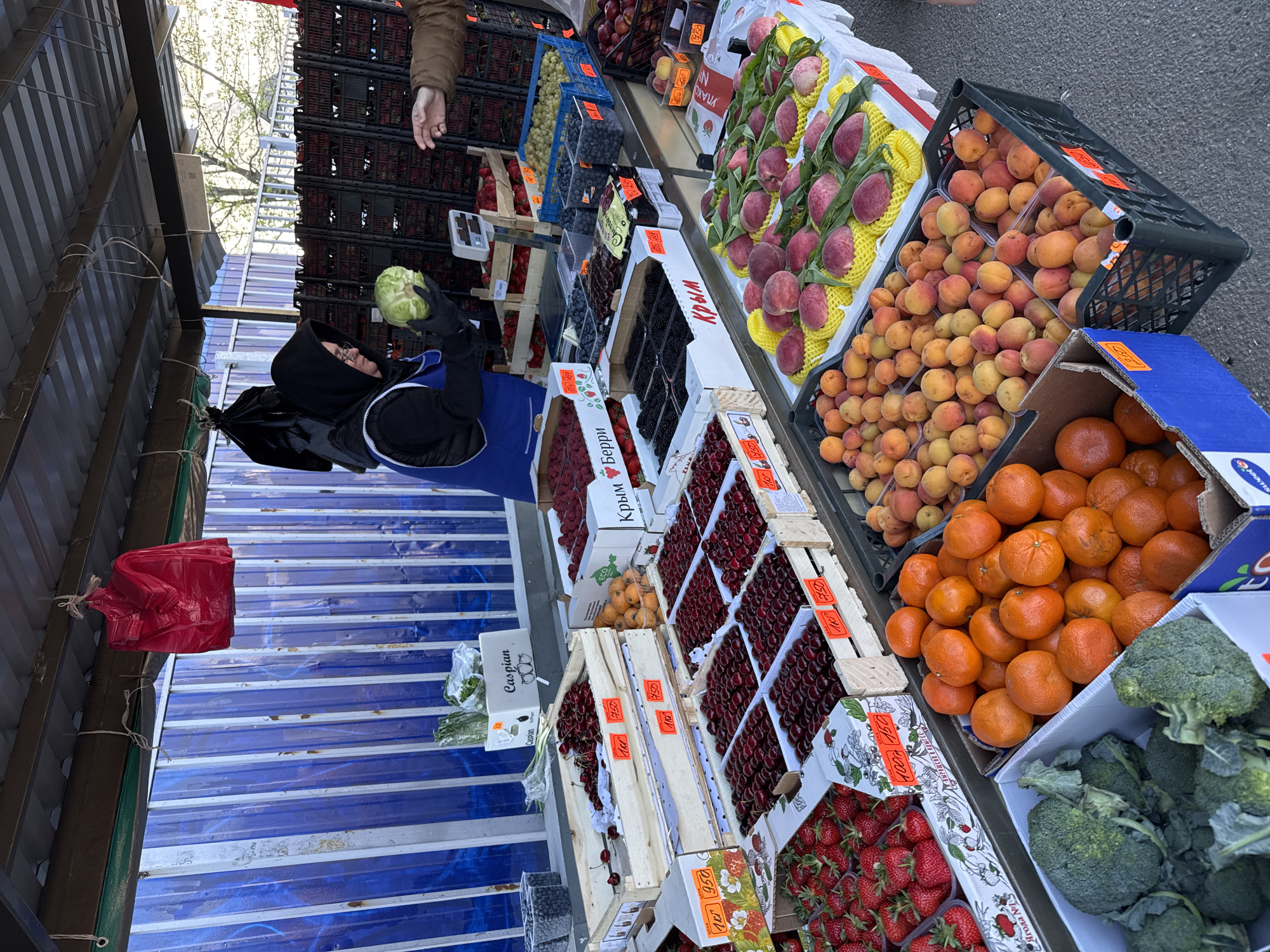 The image shows a market stall in Vsevolozhsk, Russia, full of fresh produce. The produce includes strawberries, cherries, raspberries, blueberries, blackberries, peaches, apricots, oranges, and broccoli, all displayed in crates and boxes. There is also a head of cabbage being held by a person wearing a black hooded coat and gloves. A red plastic bag hangs from the stall's roof. The stall has a blue and white striped surface under a metal roof. Prices are displayed in Russian on small signs.