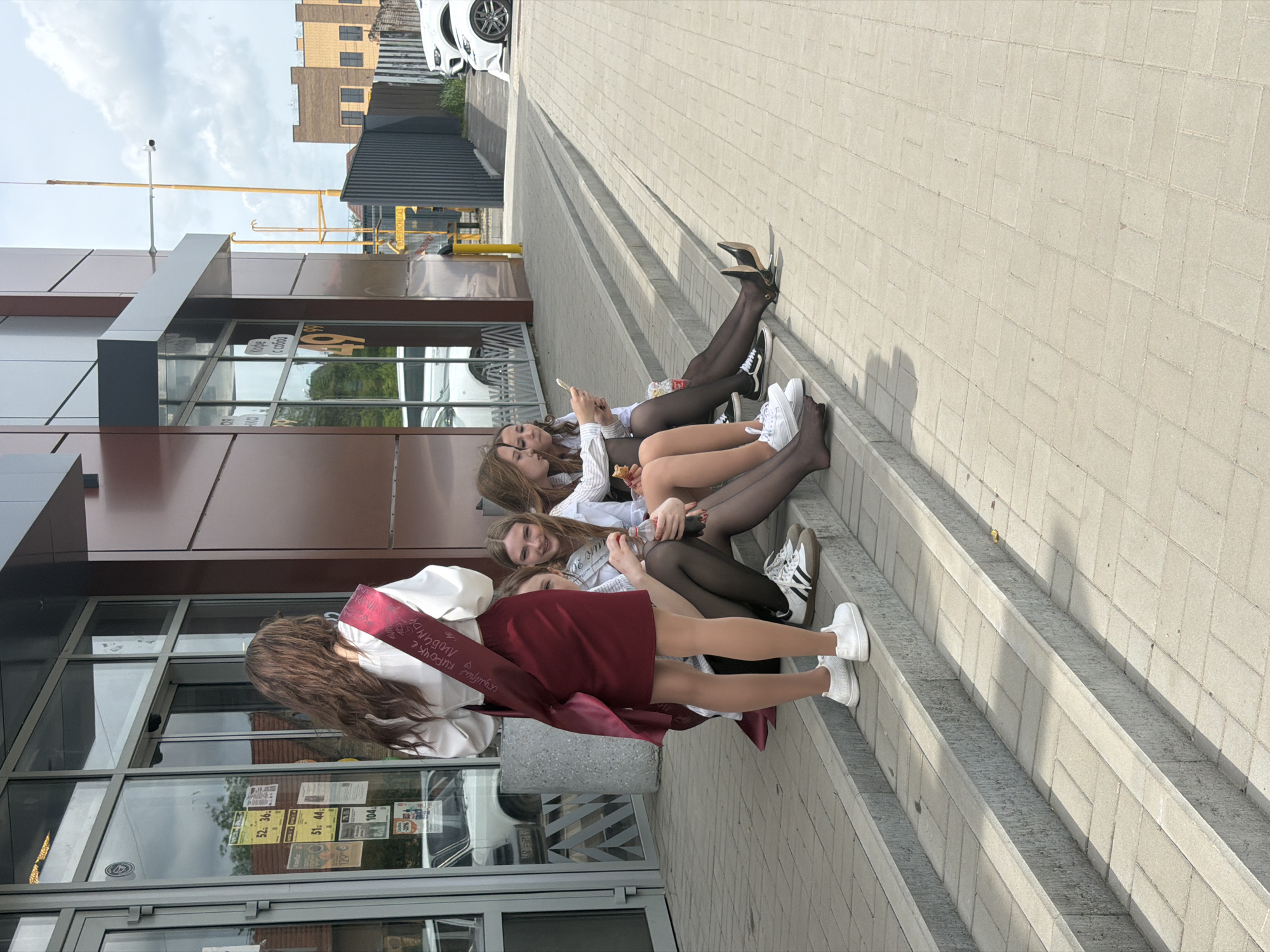 The image shows a group of four young women sitting on the steps outside a building. The woman closest to the camera is wearing a maroon sash with white text. They are likely outside a retail location in Leningradskaya, Russia. The group is on the steps of a building with glass windows. A light colored brick or stone pavement is present. A parking area with a white car is partially visible in the upper portion of the image. The women are wearing white shirts, tights, and various styles of sneakers or shoes. At least one of the women is holding a cell phone, and another is holding a drink or snack. They appear to be posing for a photograph.