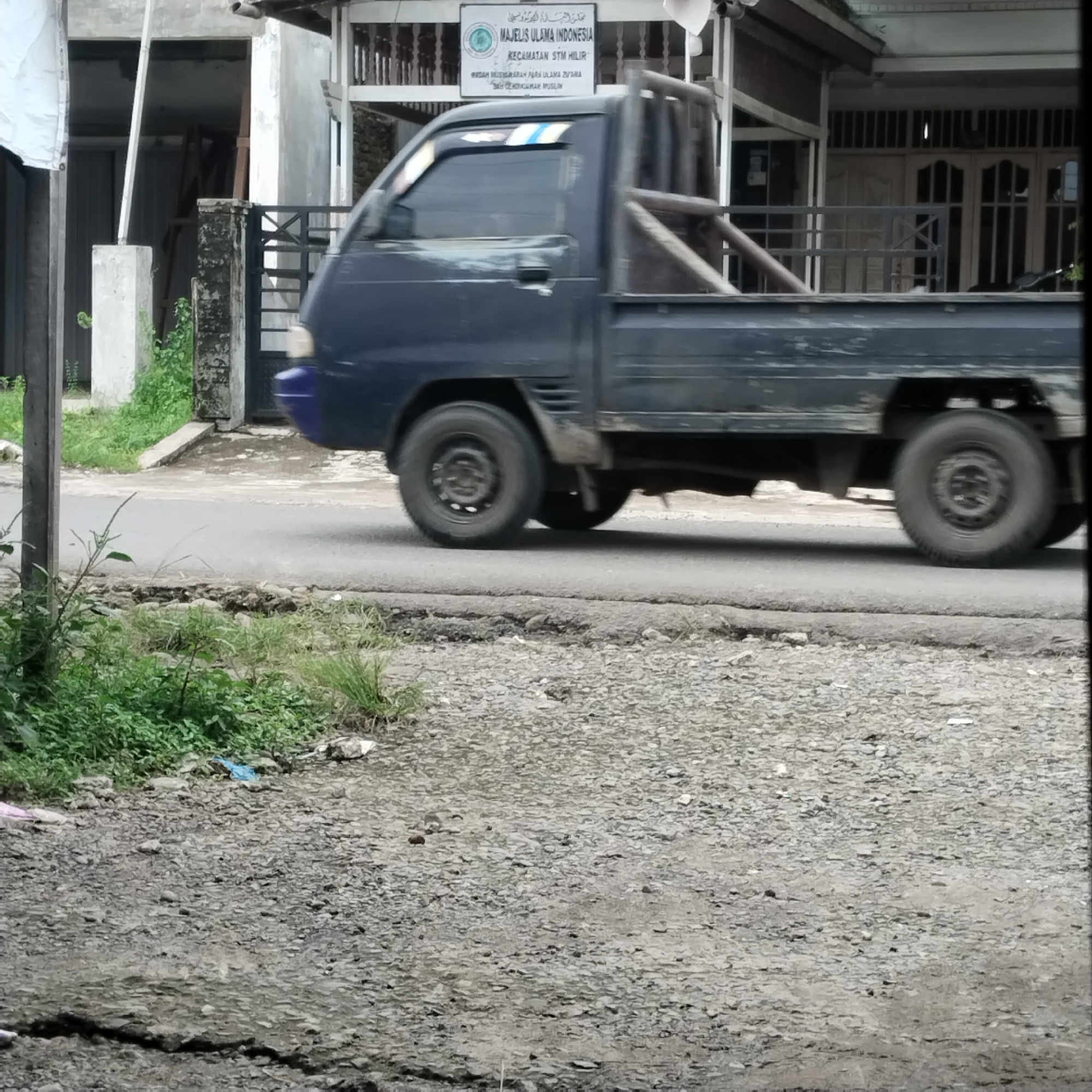 In Talun Kenas, Indonesia, a dark blue pickup truck is captured in motion on an asphalt road. The truck is moving from left to right, its tires slightly blurred due to the movement. The vehicle's side is visible, showing a utilitarian design with a metal roll cage at the rear. The foreground is a gravelly area with patches of green grass and some scattered debris, including a bright blue plastic bag. In the background, there is a building with a white facade and a dark roof structure. A sign is mounted on the building, featuring text and a circular emblem. The environment suggests a roadside scene in a moderately developed area.
