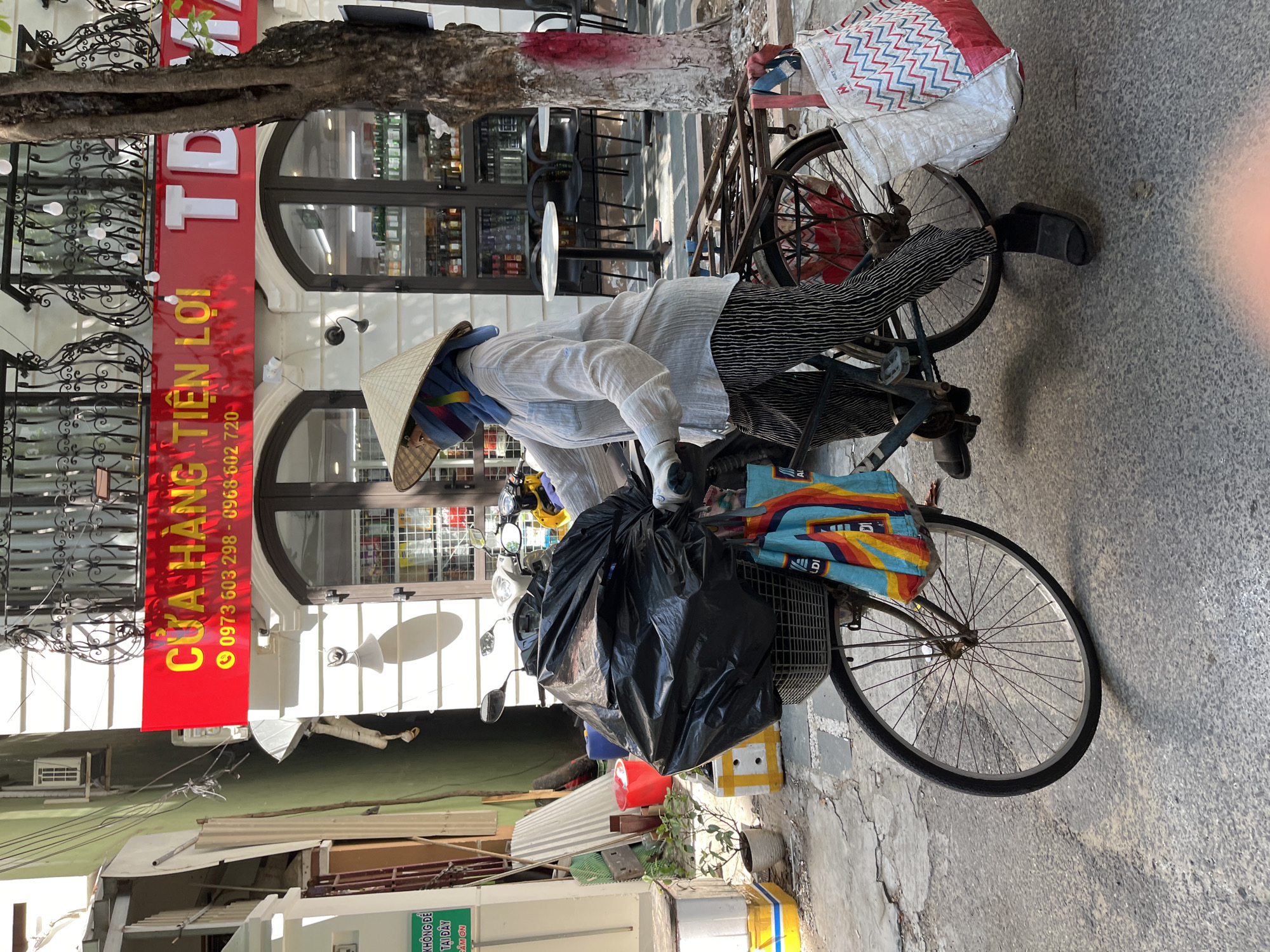 The image shows a person riding a bicycle on a street in Đà Nẵng, Vietnam. The person is wearing a traditional conical hat and a face covering, along with a light-colored long-sleeved shirt and black-and-white striped pants. The bicycle has a large black bag in the front basket and another large bag on the back. To the left of the person, there's a red sign with yellow text, including a phone number. The sign advertises a store. The street appears to be paved with light-colored stones. Behind the person and the store, there's a tree and some additional buildings.