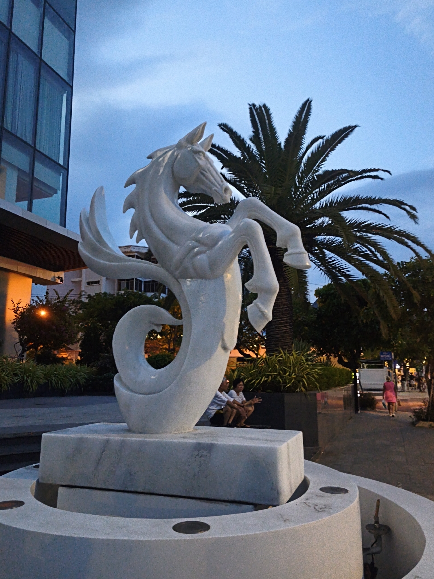 The image depicts an outdoor scene at dusk, likely in the city of Nha Trang Ward, Vietnam, given the architectural style and vegetation. The focal point is a large, white marble sculpture of a horse with a swirling, fish-like tail, mounted on a multi-tiered marble base that appears to be part of a fountain. The sculpture is positioned in front of a modern building with a glass facade, and to its right, a tall palm tree with prominent fronds is visible against the twilight sky.
In the background, a paved walkway runs alongside a street lined with trees and several businesses. Several people are present: two individuals are seated on a dark bench to the left of the sculpture, and another person, wearing a bright pink dress, is walking away from the camera down the street on the right. The lighting suggests it is evening, with streetlights and illuminated building interiors casting a warm glow on the scene. Lush green foliage surrounds the base of the sculpture and the building.