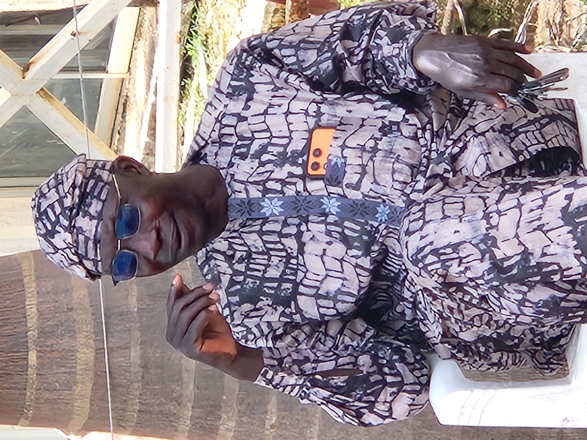 A man wearing a patterned traditional outfit and a headwrap sits in a white plastic chair. He wears blue-tinted glasses and has his left hand resting on his chin, while his right hand holds a set of keys. An orange smartphone is tucked into the breast pocket of his shirt. The background appears to be outdoors, with a palm tree trunk to the left and a wooden structure with glass panes to the right. The ground is covered with dry grass and dirt. The image is cropped to focus on the man, suggesting this scene might be in a residential area or a social gathering space in Jos, Nigeria.