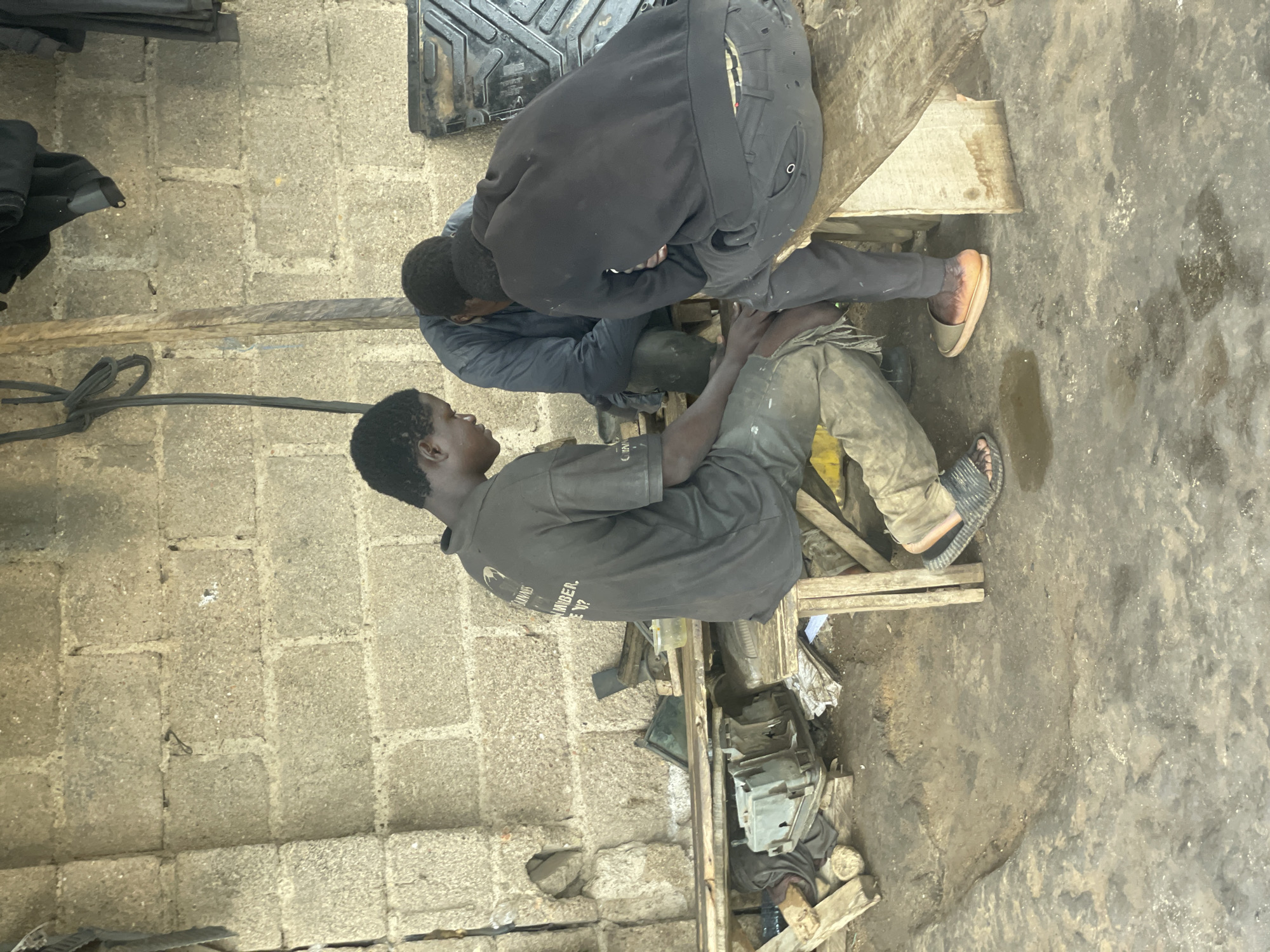 In New Stadium, Nigeria, three young men are gathered around a low wooden bench in what appears to be an open-air workshop or storage area. The background consists of a rough, unpainted cinder block wall and some hanging black fabric. The ground is dirt and appears wet in places.
The man on the left is wearing a dark, possibly grey, t-shirt with some writing on the back, and light-colored, stained trousers. He is seated and looking down at something in his lap, his hands engaged in an action. He wears dark sandals.
In the center, another young man, facing away from the camera, wears a dark blue jacket and dark trousers. He is also seated and leaning forward, his head bent.
The man on the right is viewed from the back. He wears a black hooded sweatshirt and dark trousers, with a plaid waistband visible. He is seated on the bench, facing the other two, with his legs crossed and is looking down. He wears light-colored slip-on sandals.
Scattered around the scene are various objects, including wooden planks, what appear to be discarded machine parts or electrical equipment, and a yellow plastic container. The overall impression is of a functional, but cluttered, workspace.
