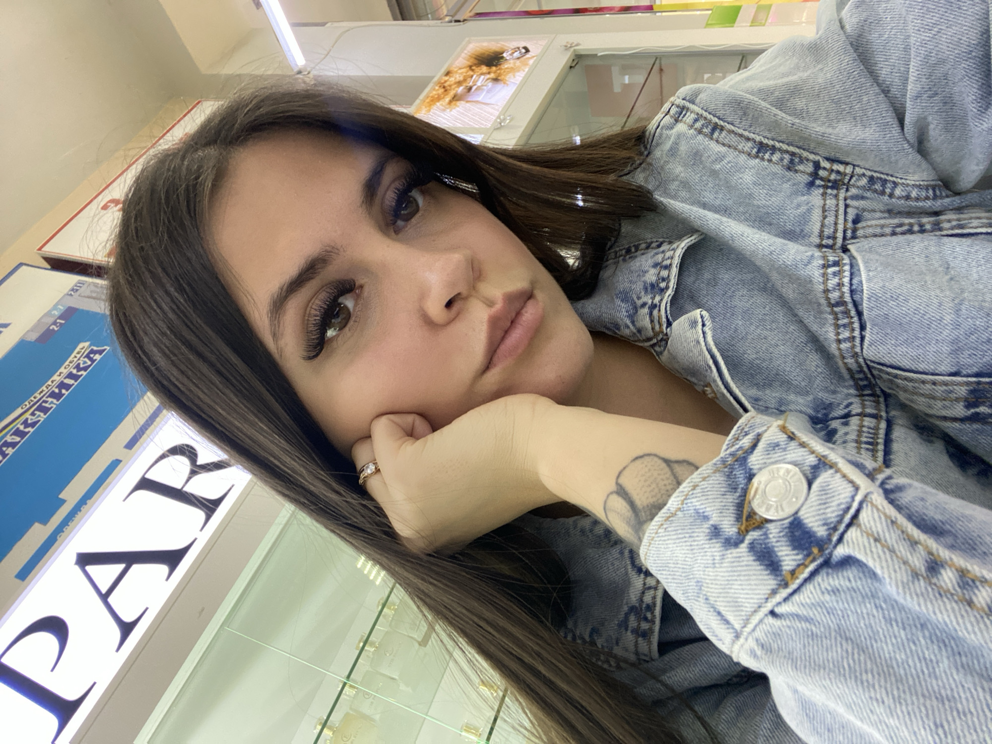 A woman with brown hair and a light blue denim jacket rests her head on her hand. She has long eyelashes and is wearing a ring. Her left arm has a visible tattoo. Behind her is a glass display case with products inside, and a sign that reads "PAR..." in large black letters. The location is likely a retail store, specifically a department store with jewelry and cosmetics. Further details of Chaikovsky, Russia cannot be inferred from the image.