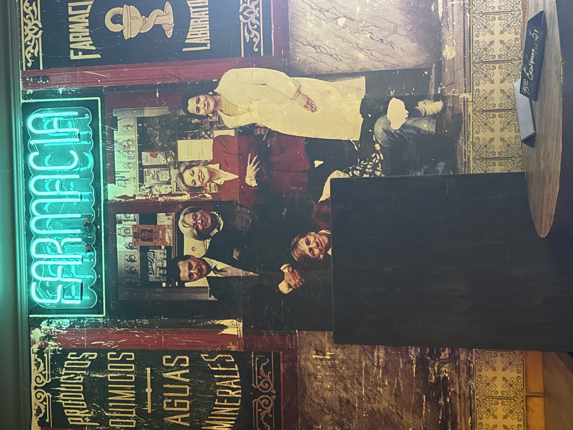 The image captures a wall in what seems to be a restaurant or bar in Moscow, Russia, decorated with vintage pharmacy posters and imagery. Prominent is a neon sign reading "Farmacia" (pharmacy), next to posters with phrases like "productos quimicos" (chemical products) and "aguas minerales" (mineral waters). There are old photographs featuring people, possibly pharmacists or individuals associated with a pharmacy setting. The wall also features vintage-style tiles. A wooden table with a menu card showing a price in rubles obscures part of the scene.