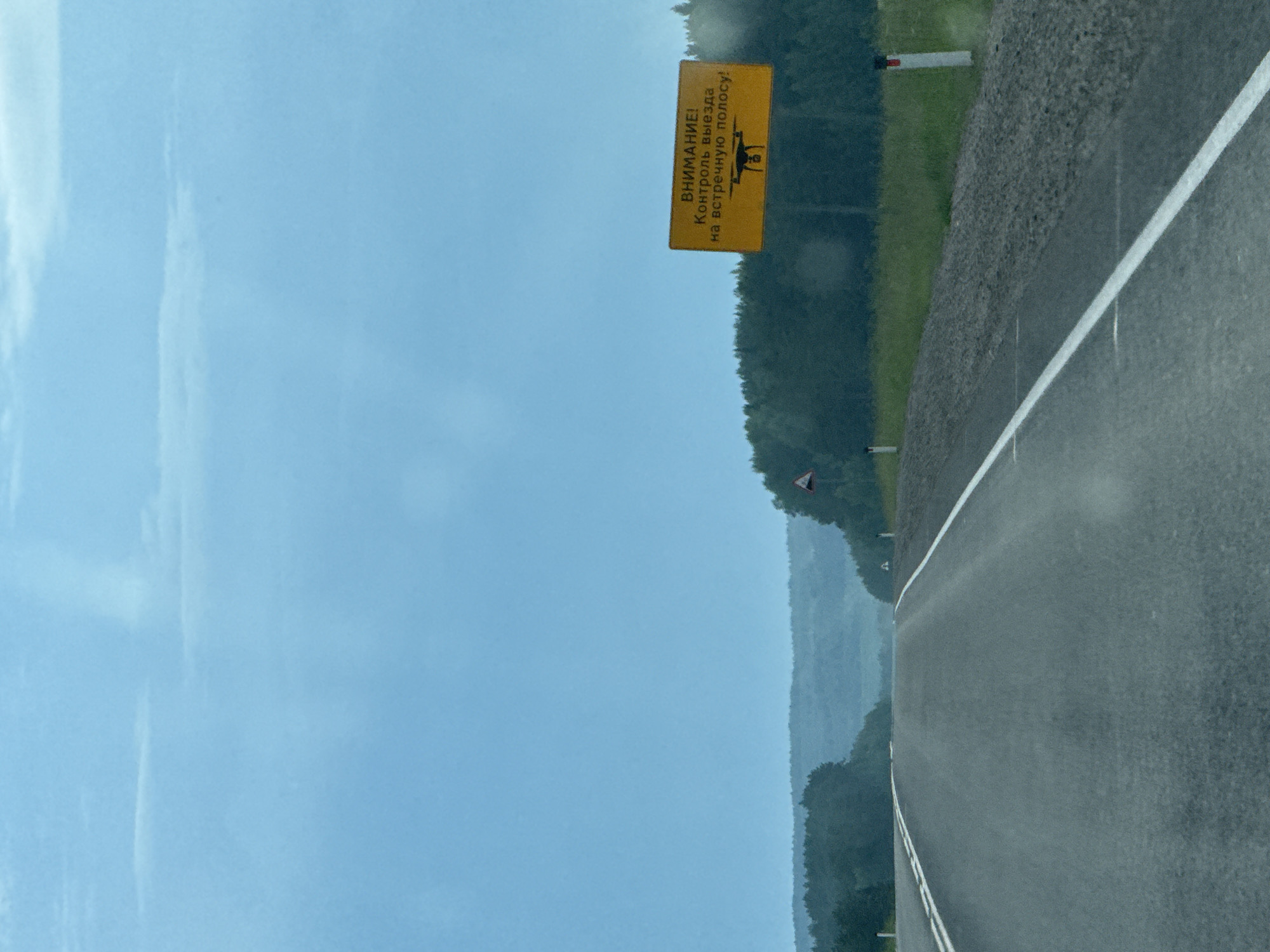 The image shows a view of a two-lane asphalt road stretching into the distance, with a lush green forest on either side. The sky is overcast with a light blue hue. On the right side of the road, a yellow rectangular sign with black lettering is prominently displayed. The sign has the text "ВНИМАНИЕ! Контроль выезда на встречную полосу!" (ATTENTION! Control of driving into the oncoming lane!) and features a black silhouette of a drone at the bottom. There are also two white road markers with red tops visible along the right edge of the road. In the distance, rolling hills covered in trees are visible. The overall scene depicts a rural highway in Karagay, Russia, with a warning sign about traffic control.