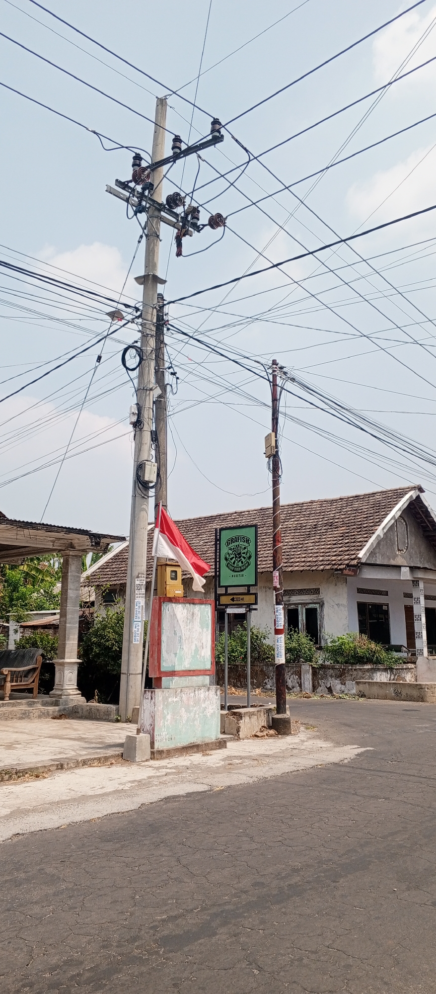 In Setren, Indonesia, a utility pole stands near a paved road, adorned with a dense network of overhead wires. Attached to the pole is a yellow electrical box and a red and white Indonesian flag. To the right of the pole, a green sign with white text and a logo reads "GRAFISH" and "-MASETAN-". Further to the right, a modest, single-story house with a tiled roof and a slightly weathered facade is visible. The house has several windows and a covered porch area. In the foreground, the road shows signs of age with visible cracks in the asphalt. To the left of the utility pole, a covered structure supported by columns stands adjacent to the road, and a wooden bench can be seen beneath it. The surrounding area is characterized by a clear sky with minimal cloud cover and vegetation.