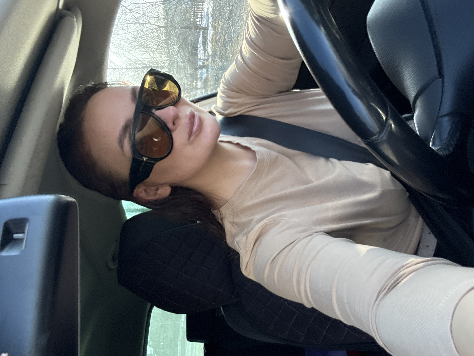 A woman wearing large, dark sunglasses and a light-colored, long-sleeved shirt sits in the driver's seat of a car. She is wearing a seatbelt. The steering wheel is visible. The location may be Chaikovsky, Russia, but there is not enough detail in the image to confirm this. Bare trees are visible through the car window.