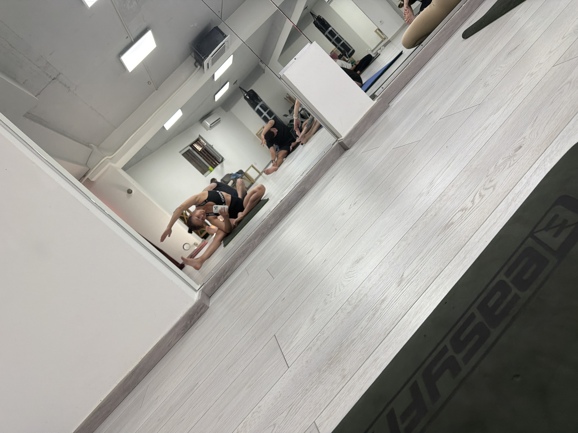 A mirror in a brightly lit gym reflects a room with several people participating in yoga or stretching exercises. One individual in the foreground is captured in a complex pose, lying on a green mat with their back arched and one arm raised, while another holds a phone. Other people are doing various exercises in the background. The gym features light-colored floors and white walls, and several punching bags are visible hanging from the ceiling in the reflected space. The lower right portion of the image shows a corner of an exercise mat with the text "EASYFIT" written in a large font. The location, Budva, Montenegro, is not discernible from the given image.