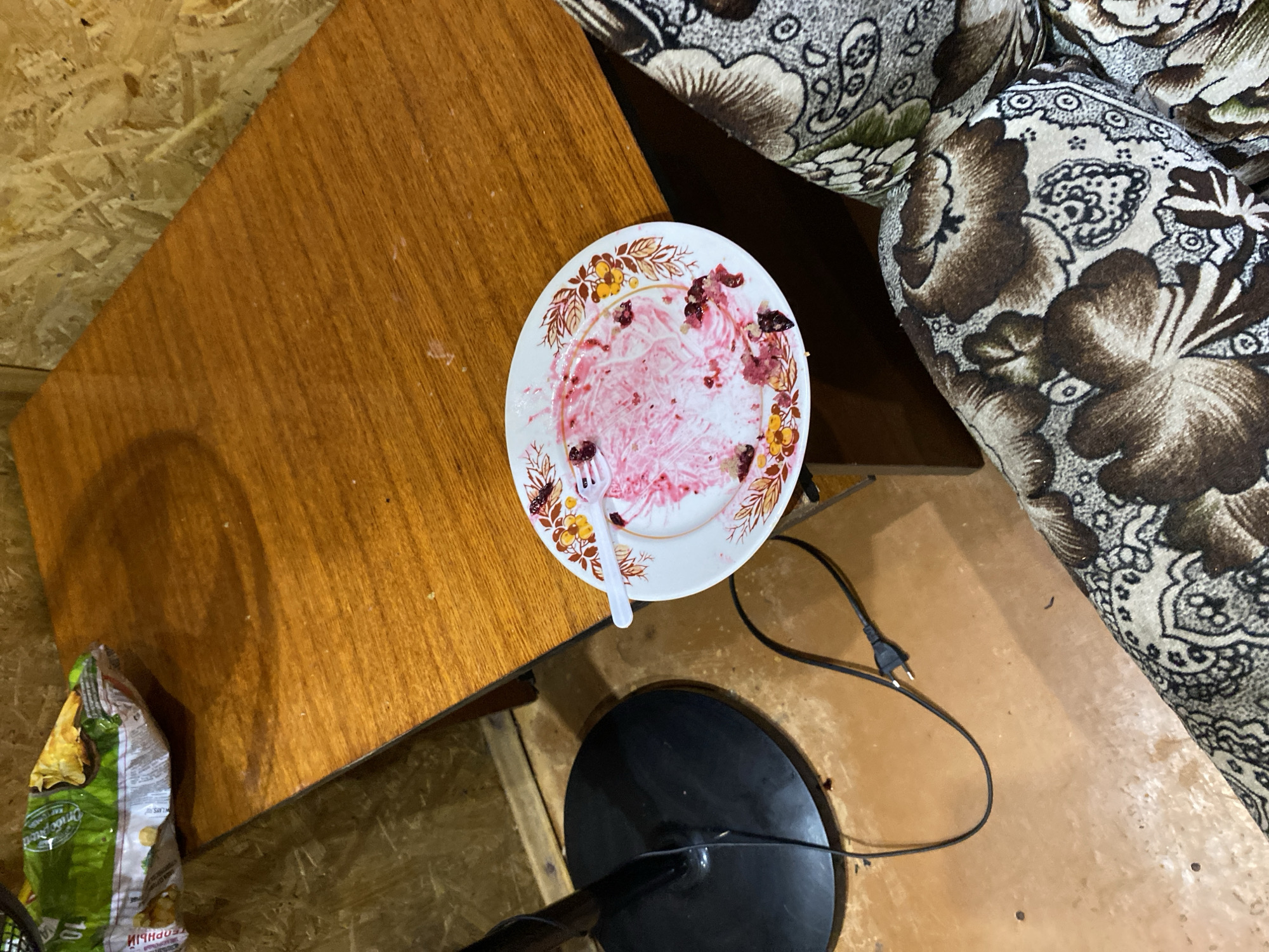 A forensic, matter-of-fact description of the scene:
The image depicts a close-up, high-angle view of a cluttered, indoor environment, likely a residential living space in Zima, Russia. The primary focus is a wooden table with a glossy, orange-brown finish, showing signs of wear and tear with faint scratches and dust. A white plate with a floral rim, containing remnants of a pink dessert, possibly cake or ice cream, and a single white plastic fork, rests on the table's surface. To the left of the table, the black base of a floor lamp is visible, connected to a black electrical cord that runs across the floor. In the upper left corner, a crumpled green and yellow plastic bag, possibly containing snacks, is partially visible on the table. The background consists of light-colored, textured walls, possibly made of particleboard or wood paneling. In the lower right portion of the image, a portion of a sofa or armchair is visible, upholstered in a brown and cream floral fabric. The lighting appears to be dim, creating shadows and highlighting the textures of the objects. There are no people or animals present in the image, nor any indication of recent activity beyond the used plate and fork.