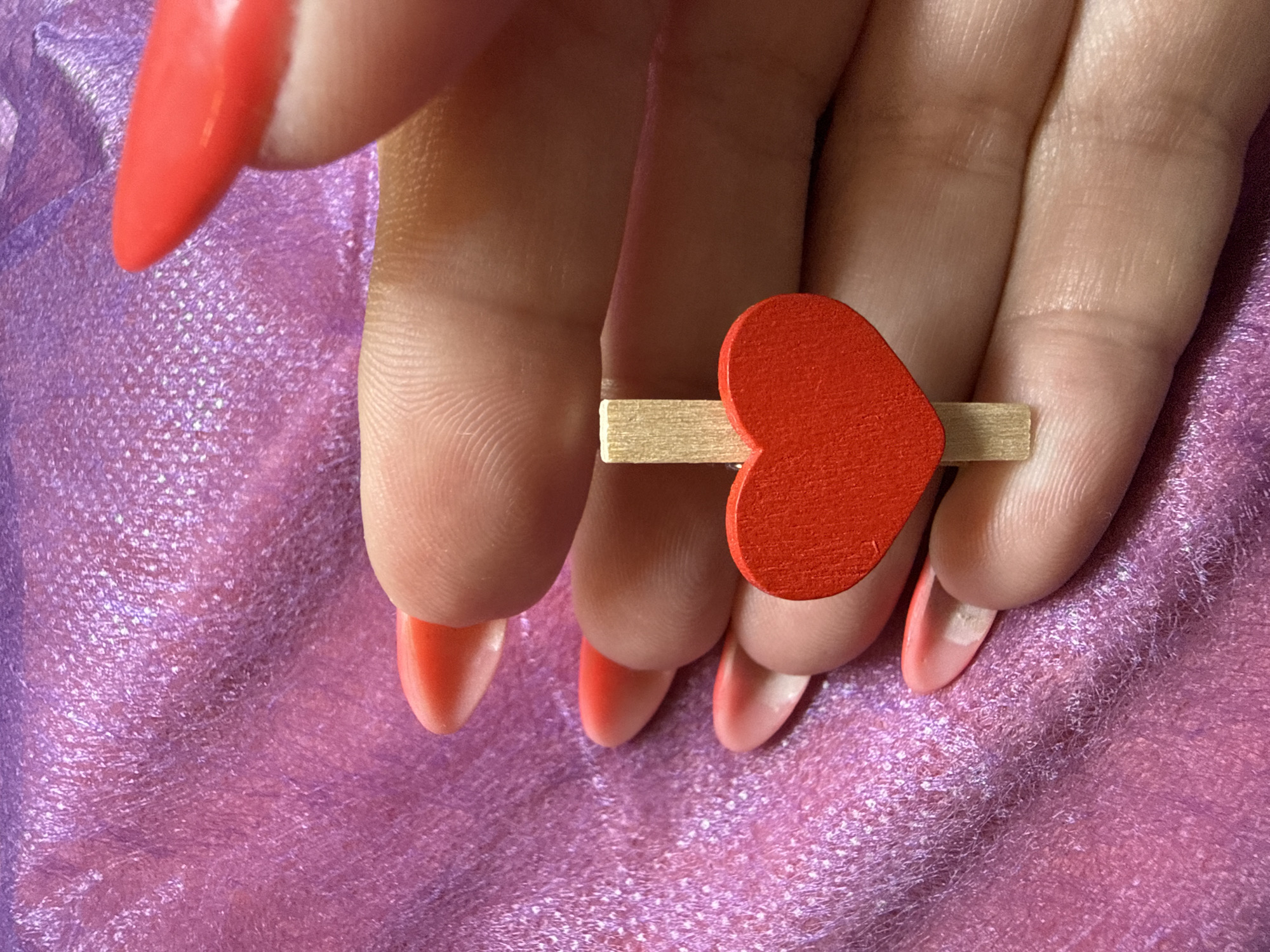 The image displays a close-up, overhead shot of a person's hand holding a small, red, heart-shaped clothespin against a textured, shimmering, pinkish-purple fabric. The hand has long, oval-shaped nails painted in a gradient of pink and orange. The fabric appears to be a textured material, possibly silk or satin, with a subtle sheen. The focus is on the clothespin and the hand, with the fabric serving as a soft background. The image does not contain any elements that would allow for identification of a specific location in Pamplona, Spain.