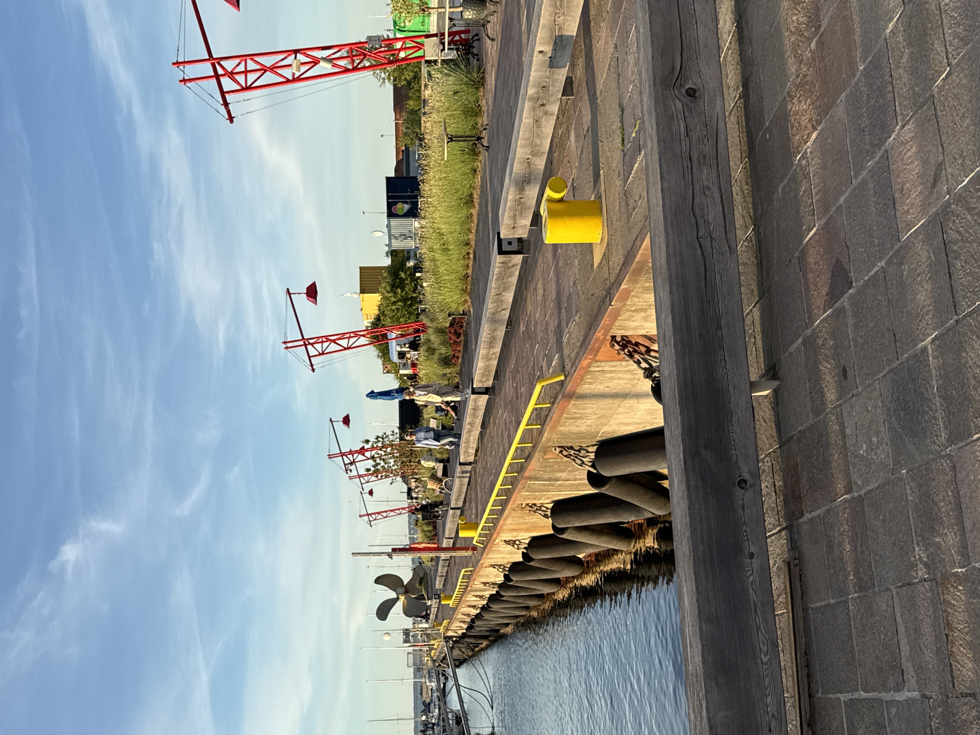 This image depicts a waterfront scene in Tallinn, Estonia, likely during the late afternoon given the long shadows and warm light. In the foreground, a paved walkway made of dark stone tiles runs horizontally across the bottom of the frame. A thick, weathered wooden beam acts as a railing or barrier, obscuring a portion of the walkway and the concrete edge of the pier.
To the left, a body of water, possibly a harbor or canal, is visible. The water is calm and reflects the sky and surrounding structures. Along the edge of the concrete pier, a row of large, black fenders hangs down, attached by heavy chains. A yellow bollard, a short, stout post used for mooring, stands on the pier.
The pier itself is a long, narrow walkway. A bright yellow railing runs along the edge of the pier, separating it from the water and fenders. In the mid-ground, several people are walking along the pier. Two women are most prominent, one in a light-colored blazer and another in a light suit, walking towards the viewer. Behind them, other individuals are visible in the distance, some sitting on benches or walking.
Further down the pier, several striking red metal structures rise into the sky. These appear to be decorative elements or possibly functional structures with lights or lamps attached. Behind these structures, there is a line of green grass and vegetation. In the background, various buildings and shipping containers are visible, suggesting an urban or industrial waterfront area. The sky above is a clear blue with scattered, wispy clouds. A large, dark propeller is mounted on a pedestal on the left side of the pier.