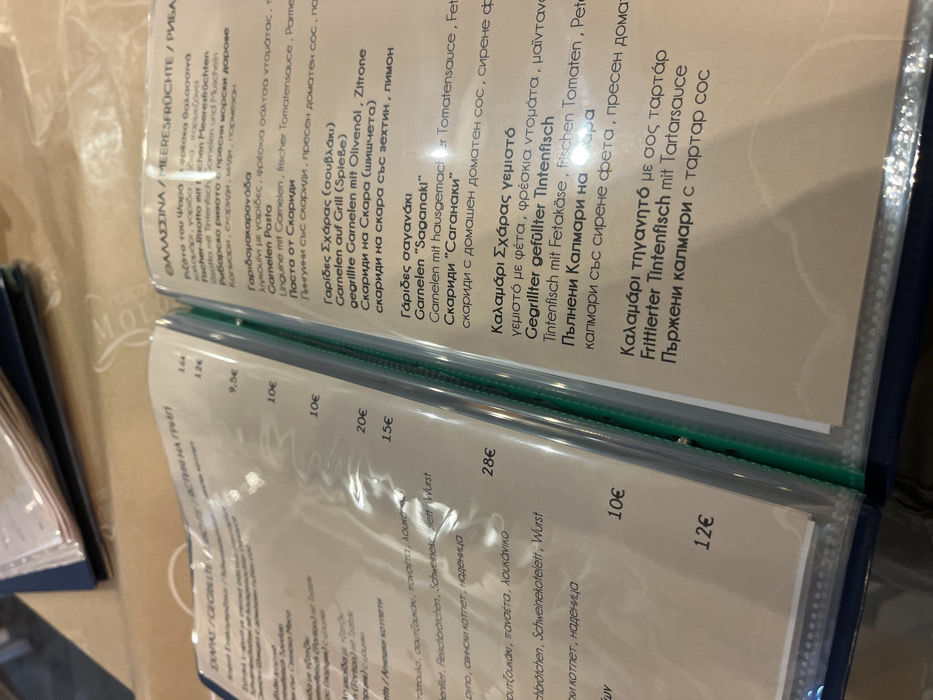 The image shows a page from a menu in Limenaria, Greece, located at the coordinates 40.628, 24.575. The page features a list of seafood dishes written in Greek and German. The menu is printed on white paper with a blue cover. Some of the dishes include: Garides Saganaki (Shrimp Saganaki), Kalamari Sxaras yeimosto (Stuffed Calamari), and Frittierter Tintenfisch mit Tartarsauce (Fried Squid with Tartar Sauce). The prices are listed in Euros. The page is open to reveal the next page, which is also part of the menu. The menu is laying on a table, likely in a restaurant. The image does not show any people.