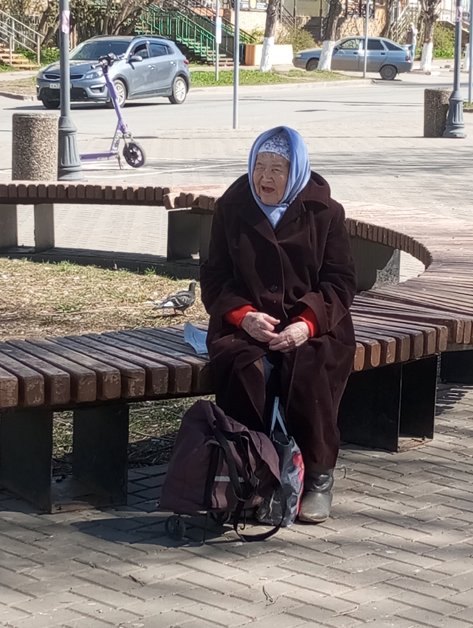 An elderly woman wearing a dark brown overcoat and a blue headscarf is sitting on a curved wooden bench in Pervomaysky, Russia. She has red sleeves peeking out from under the coat. A rolling bag is next to her feet, which are clad in grey boots. A pigeon is on the ground near the bench. In the background, cars pass by on a street, and there is a parked purple scooter. The setting appears to be a public park or sidewalk area with trees, streetlights, and other benches.
