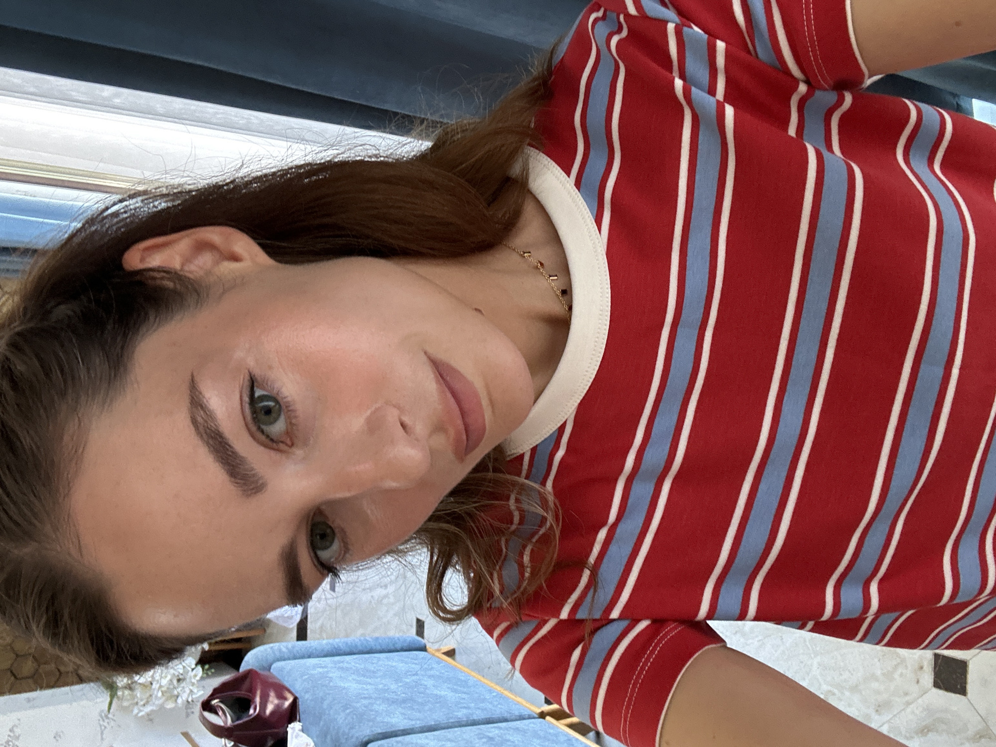 A close-up, rotated portrait image shows a young woman indoors. She has light to medium brown hair, green eyes, and well-defined eyebrows. Her expression is neutral or slightly pleasant. She is wearing a red t-shirt with vertical light blue and white stripes and a white collar. A delicate gold-colored necklace with small dark charms is visible around her neck.
The background suggests an indoor setting, possibly a home or lounge area. To her left (when the image is correctly oriented), a light blue upholstered piece of furniture is visible. Further left, there are white decorative items, possibly flowers, and a dark red object that could be a bag. A tiled floor with light and dark squares is partially visible on the right. Natural light streams in from a window with blinds, indicating it is daytime. The upper part of the background features a dark, horizontal structure.
The woman appears to be posing for the photograph. There is no visible text in the image. The location context provided is Makhachkala, Russia.